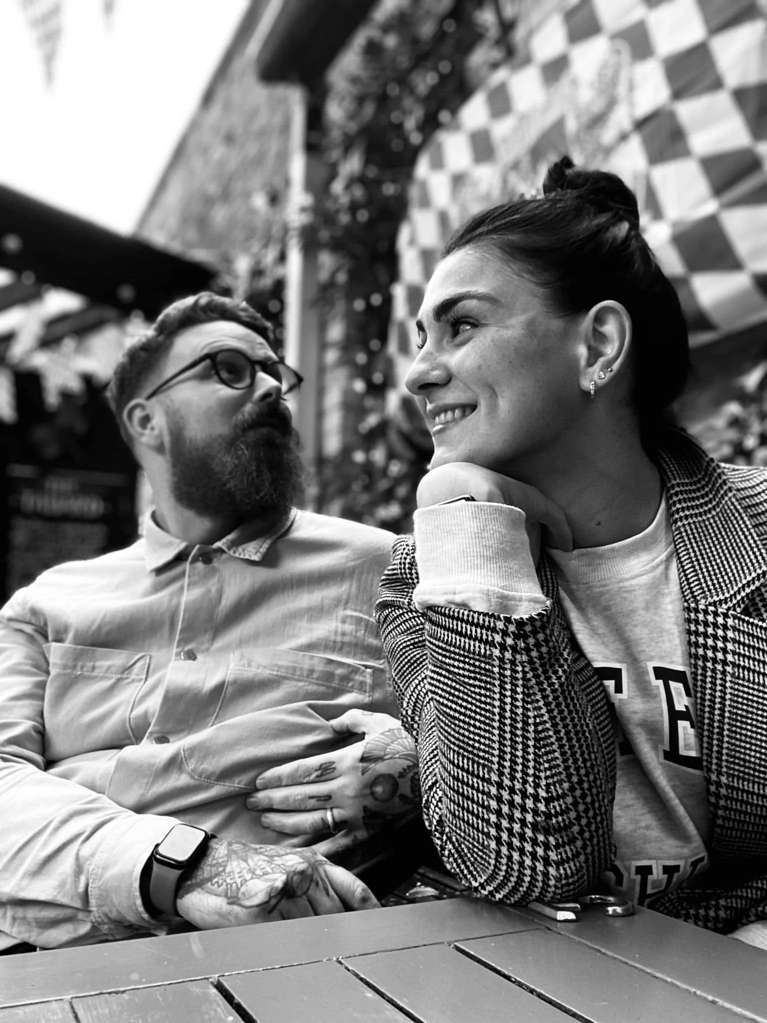 A black-and-white photo of a man with glasses and a beard looking at a woman smiling, outdoors at a cafe or patio, with brick and plant decor in the background.