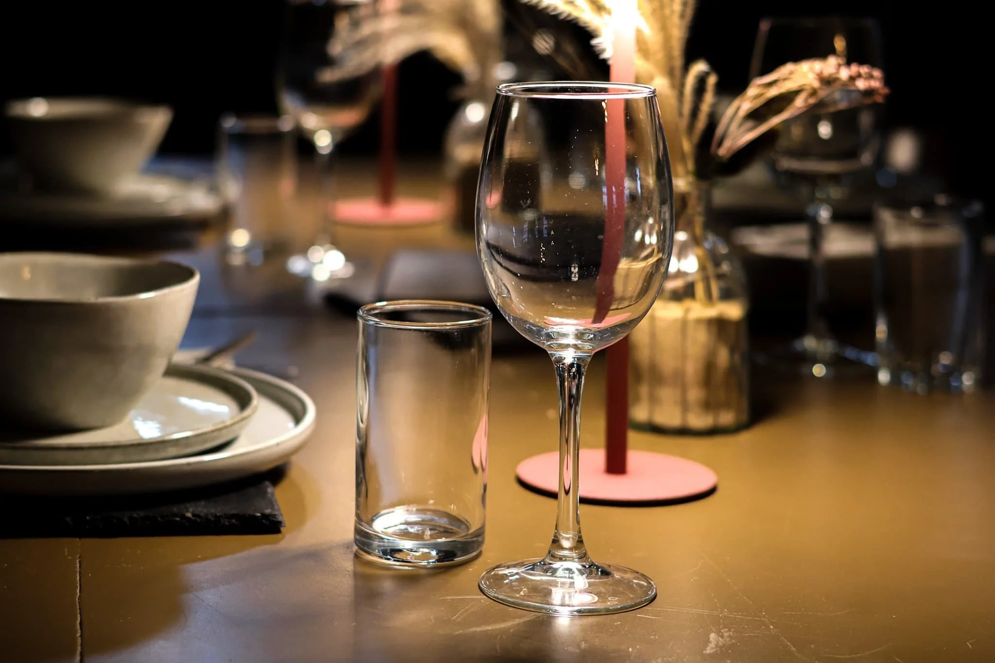 Empty wine glass and small glass on a table with dinnerware and decorative candles in the background.