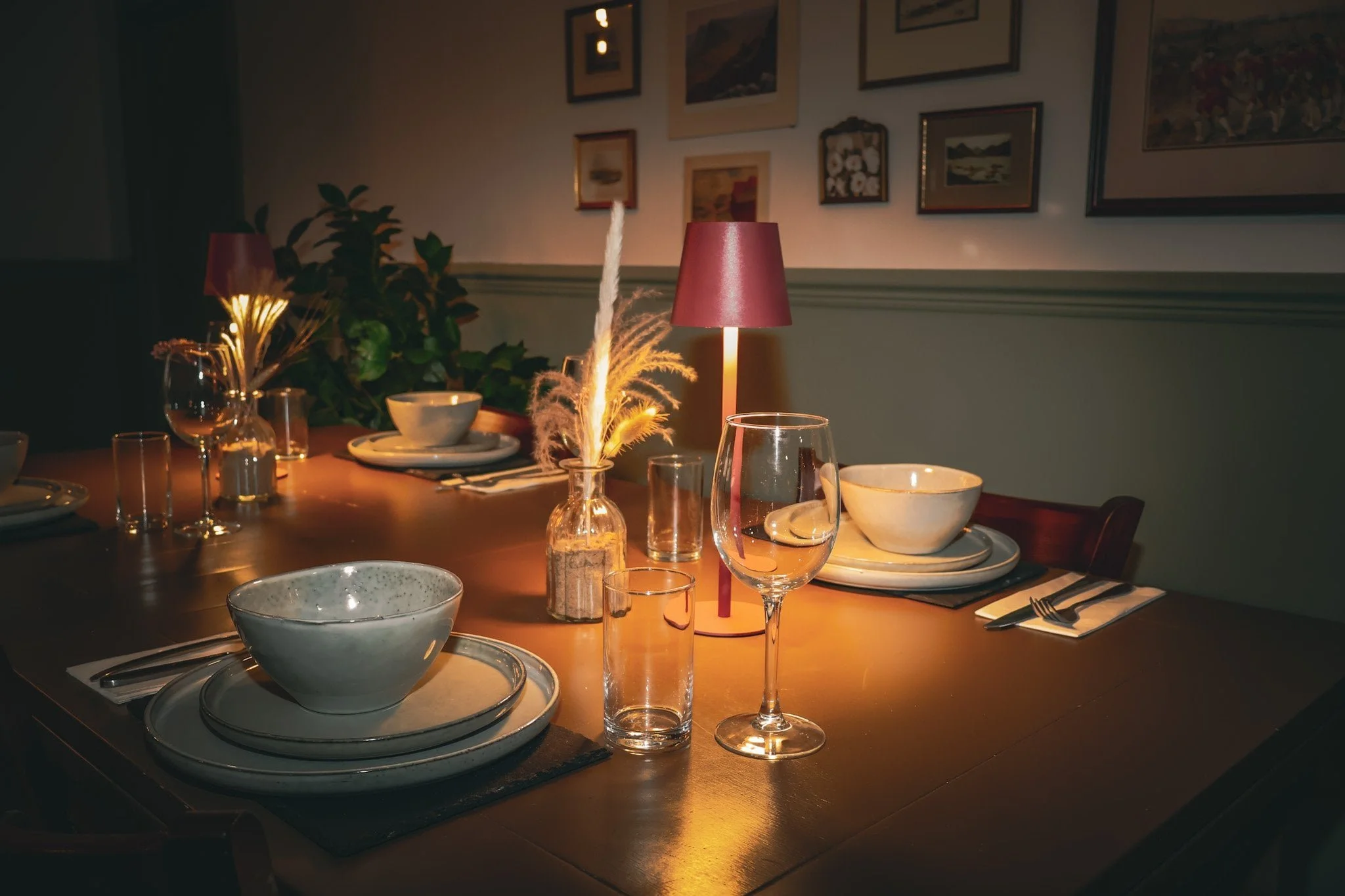 A dining table set for a meal with bowls, plates, glasses, and cutlery, illuminated by a pink lamp and decorated with a small vase of pampas grass, with framed artwork on the wall in the background.