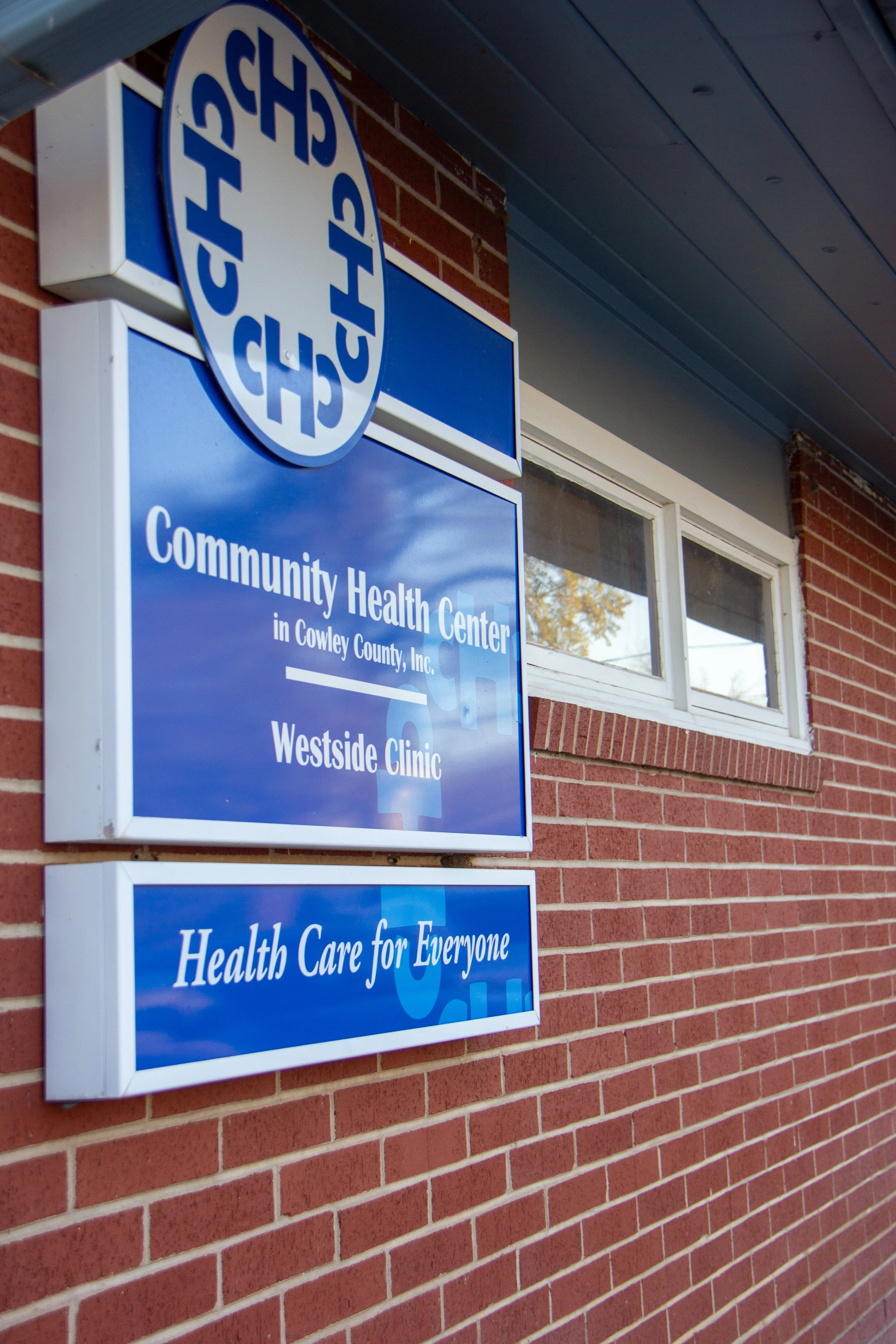 Sign on a brick building with blue and white colors, displaying the name and services of the Community Health Center in Cowley County, Inc., including the Westside Clinic, with a window visible above the sign.