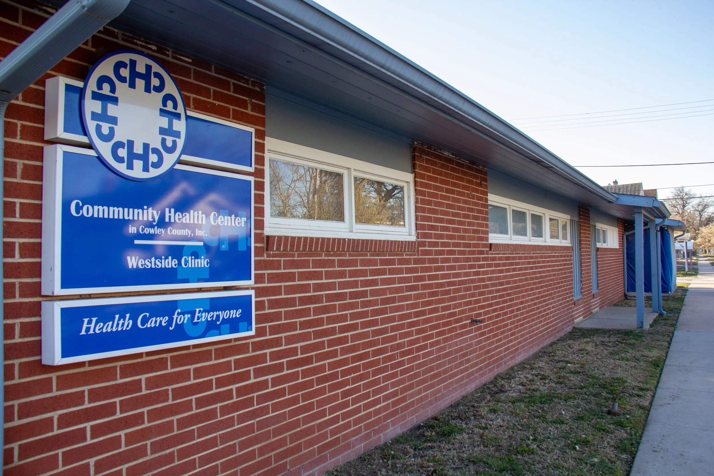 Exterior view of a brick community health center building with a blue sign reading "Community Health Center in Cowley County, Inc., Westside Clinic" and slogan "Health Care for Everyone."
