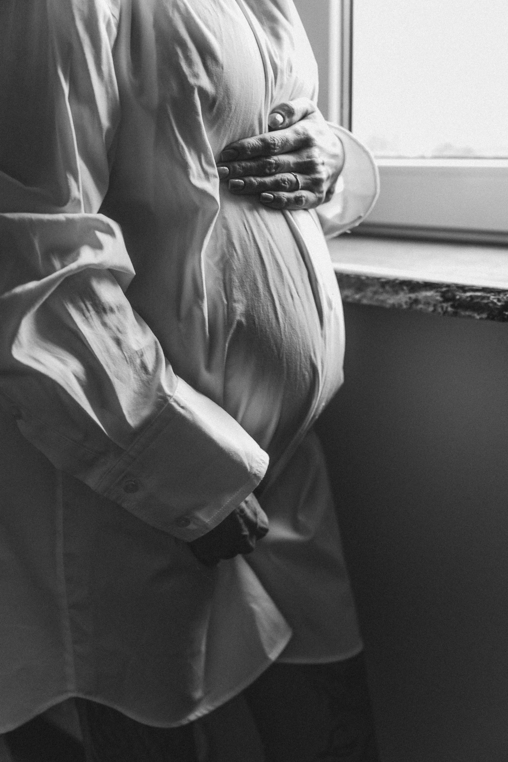A pregnant woman wearing a button-up shirt and pants, standing by a window with her hands resting on her belly.
