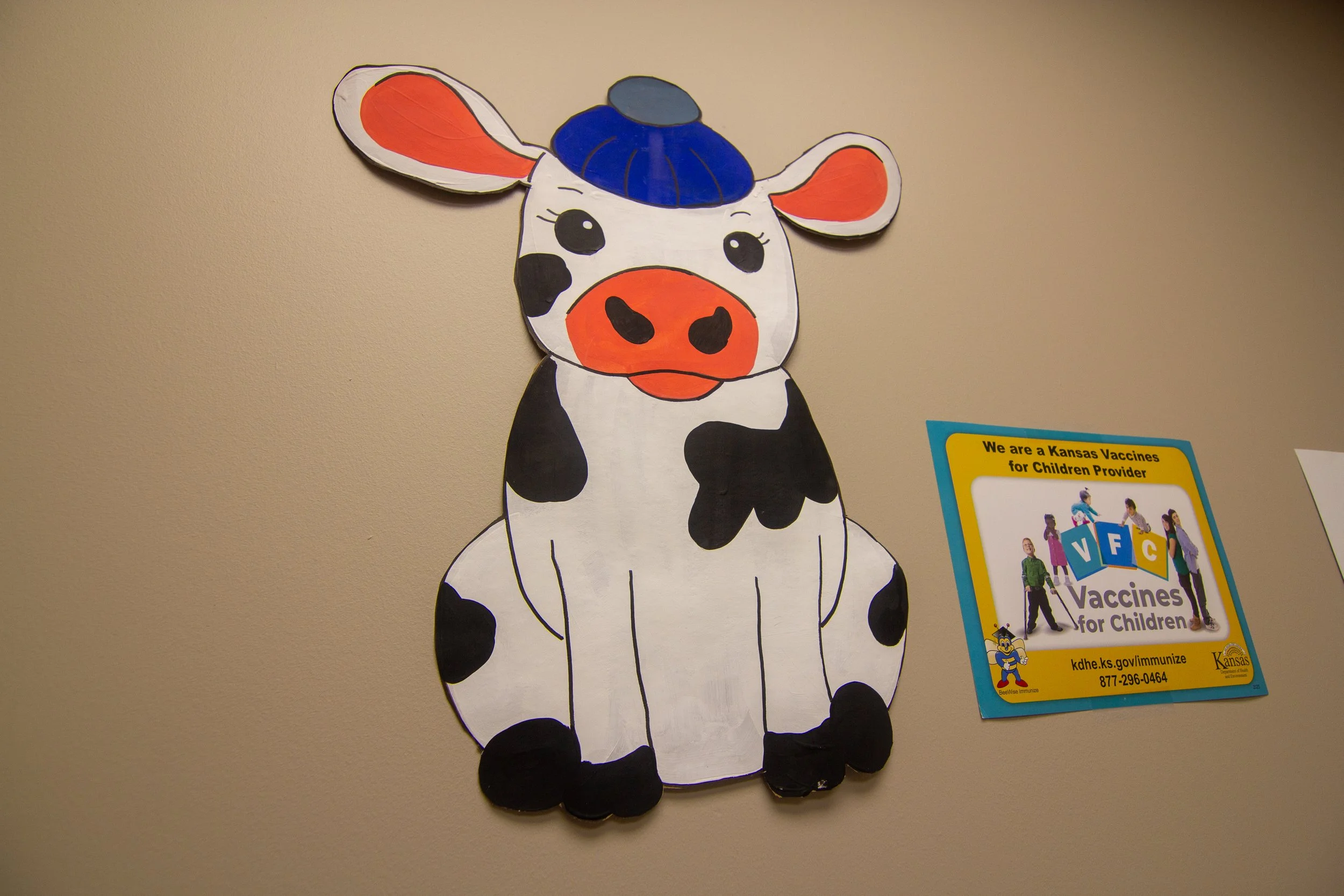 A paper craft of a cartoon cow with black and white spots, a big orange snout, and large ears, wearing a blue cap, on a beige wall. Next to it is a colorful sign promoting childhood vaccines.