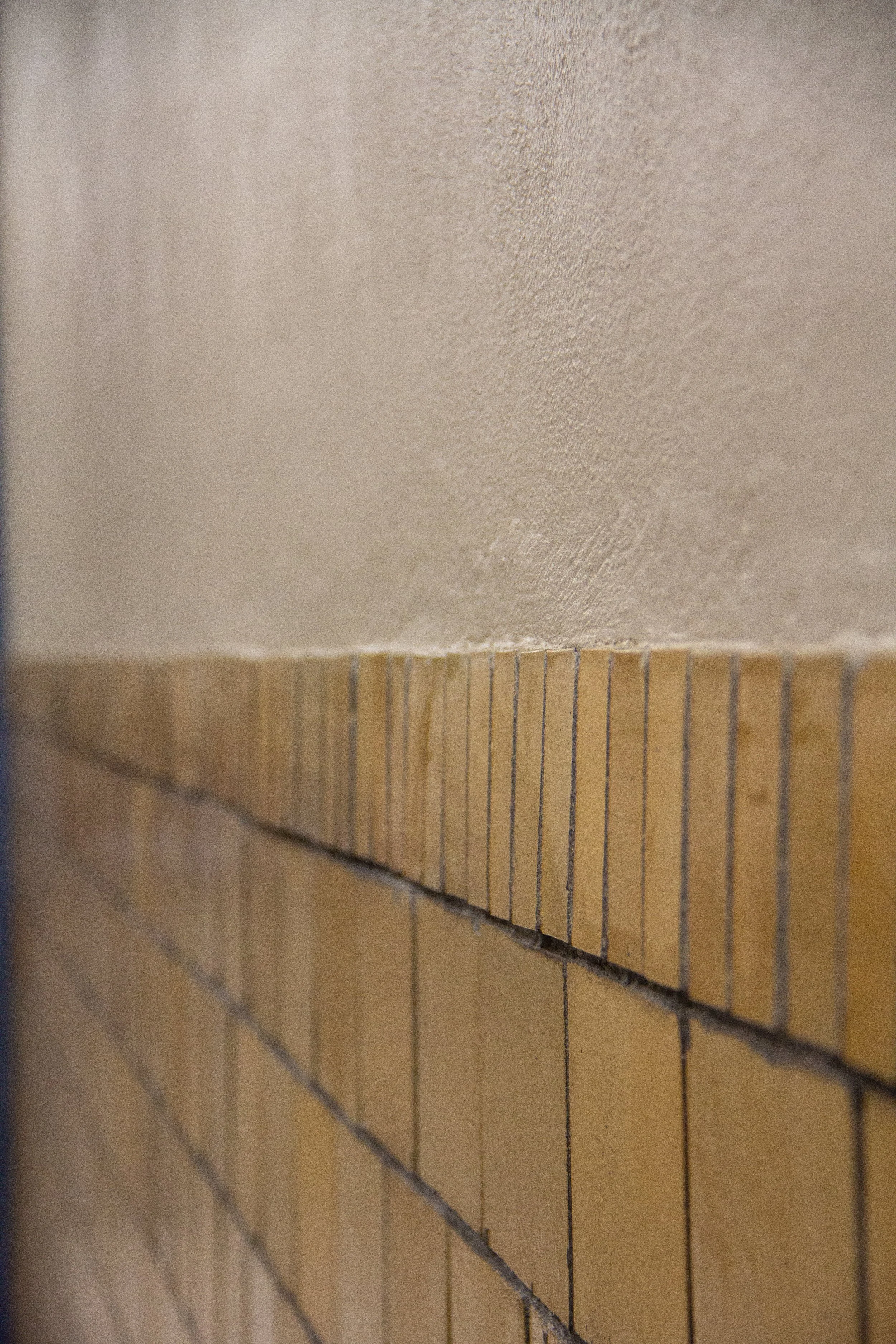 Close-up view of a wall with a textured beige upper section and a lower section made of small, rectangular yellowish tiles arranged in a grid pattern.