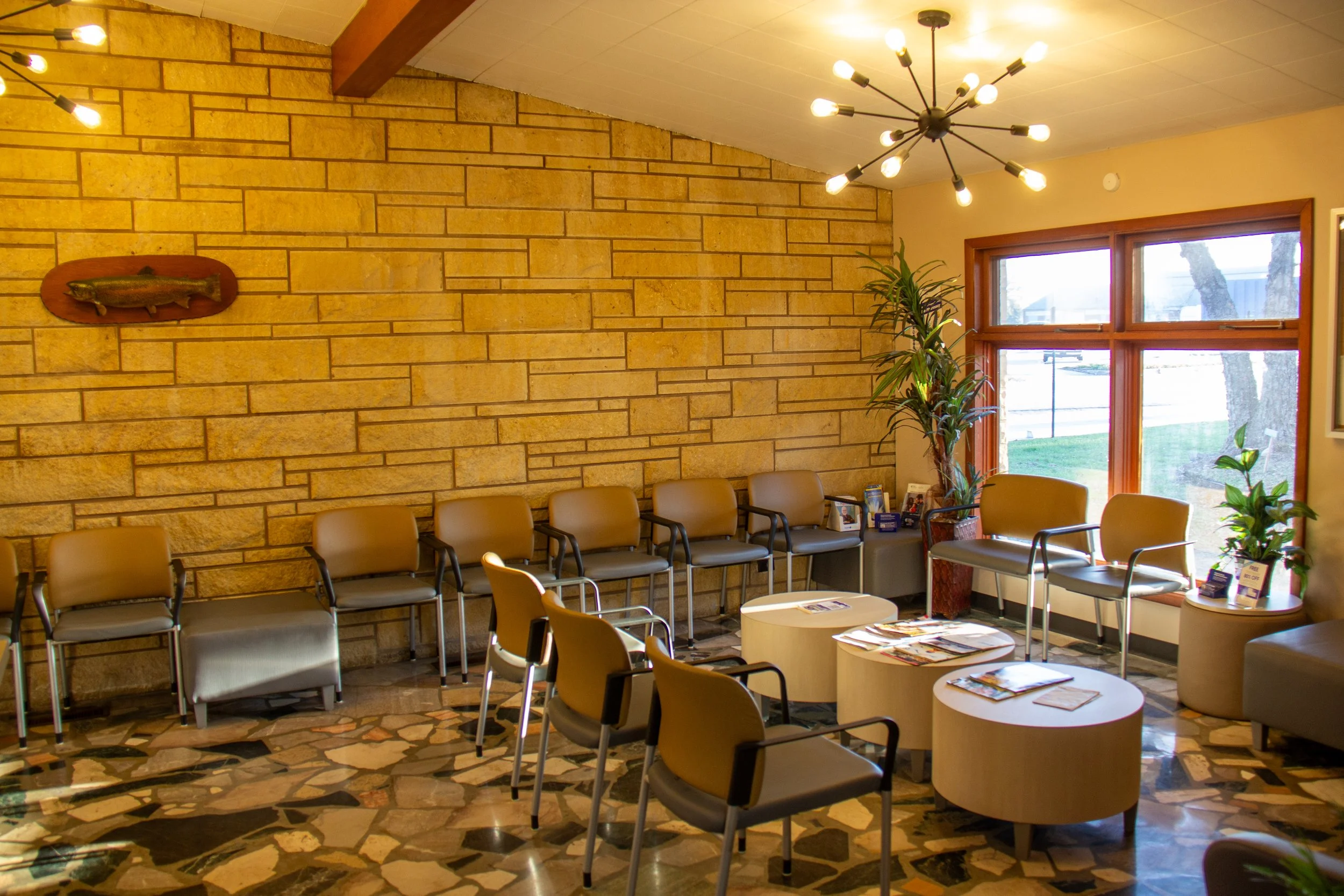 A waiting room with beige chairs arranged along the walls and around small round tables, large windows with wooden frames, potted plants, magazines on the tables, a stone accent wall, and a modern chandelier ceiling light.