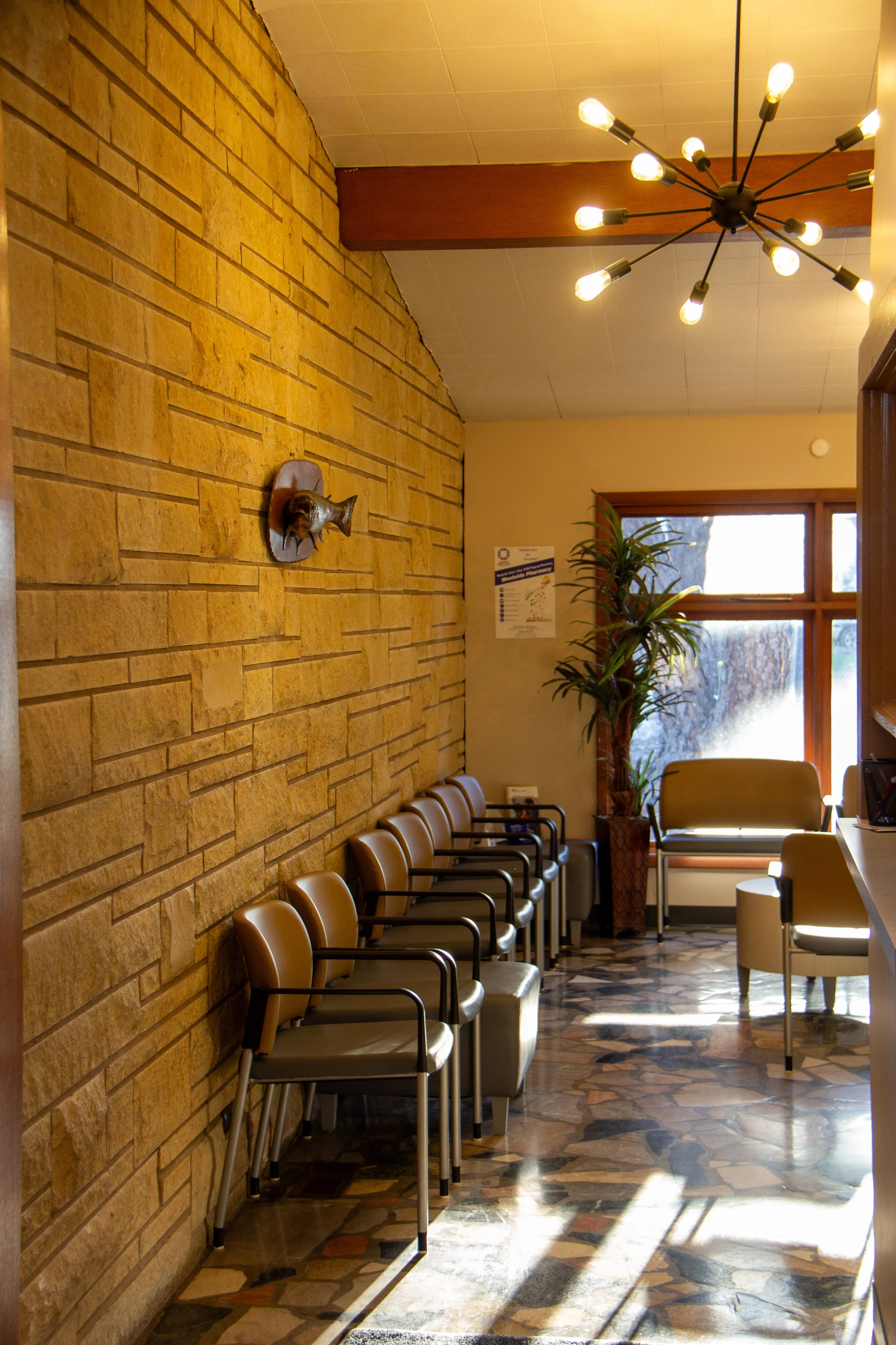 Empty waiting room with beige chairs, a potted plant, a window with sunlight, a stone-patterned floor, and a modern ceiling light fixture.