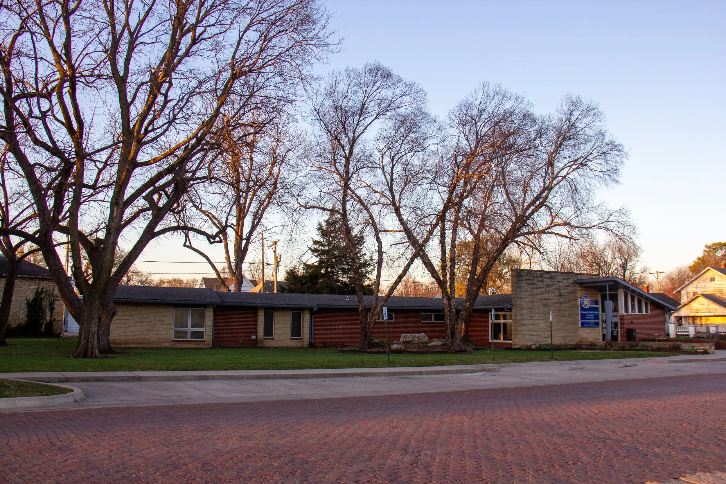 A single-story brick building with large windows, surrounded by leafless trees, with a sidewalk and a brick-paved street in front, during sunrise or sunset.