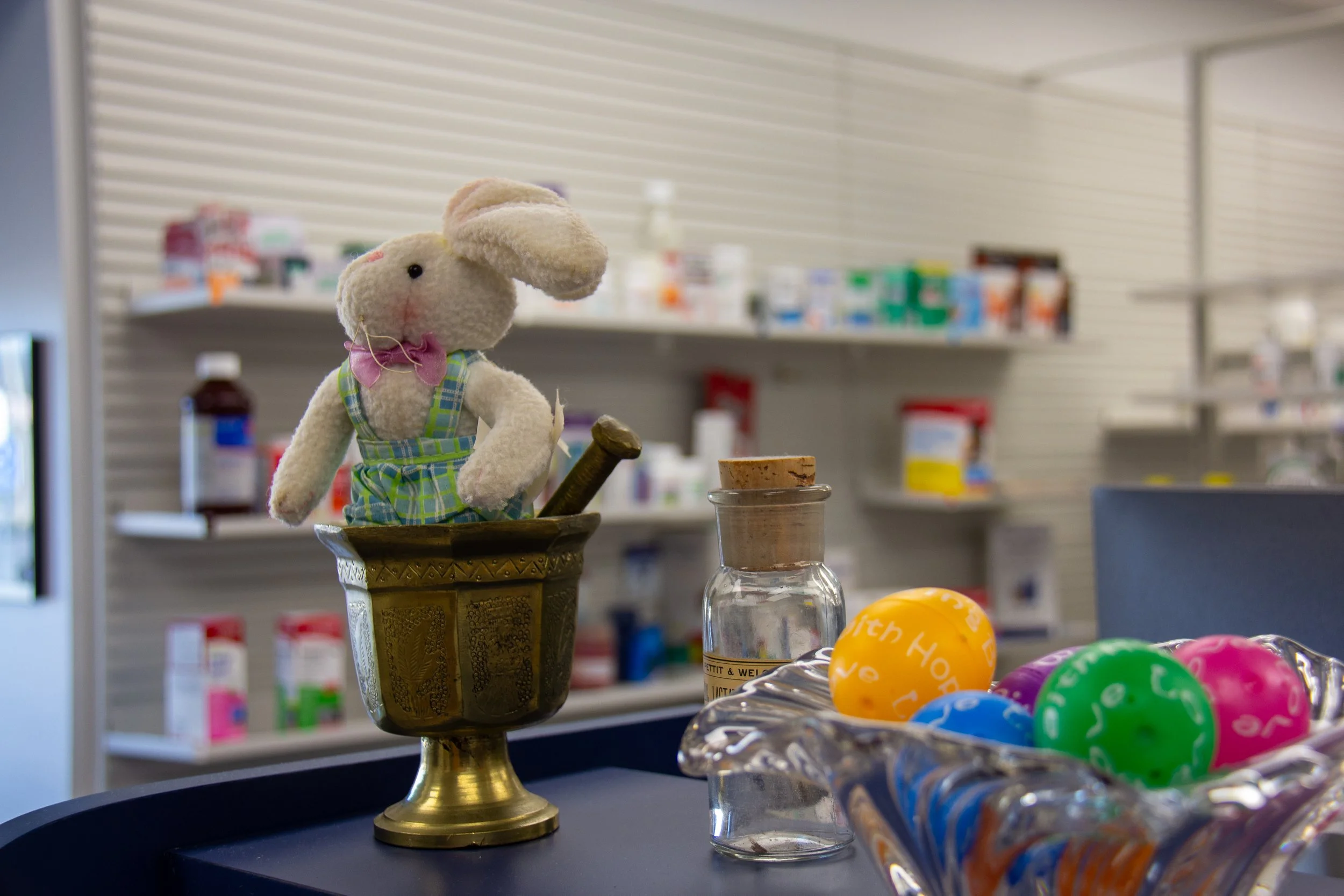 A plush bunny toy with a pink bow around its neck, dressed in a plaid outfit, standing inside a decorative gold pedestal container with painted details, on a counter in a pharmacy or store. Next to the bunny are colorful plastic eggs in a clear plast