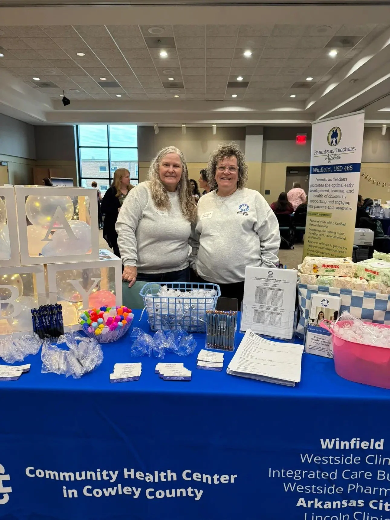 Two women standing behind a table at a community health event, with informational pamphlets, pens, and promotional items on display. A banner in front of them reads 'Community Health Center in Cowley County.'