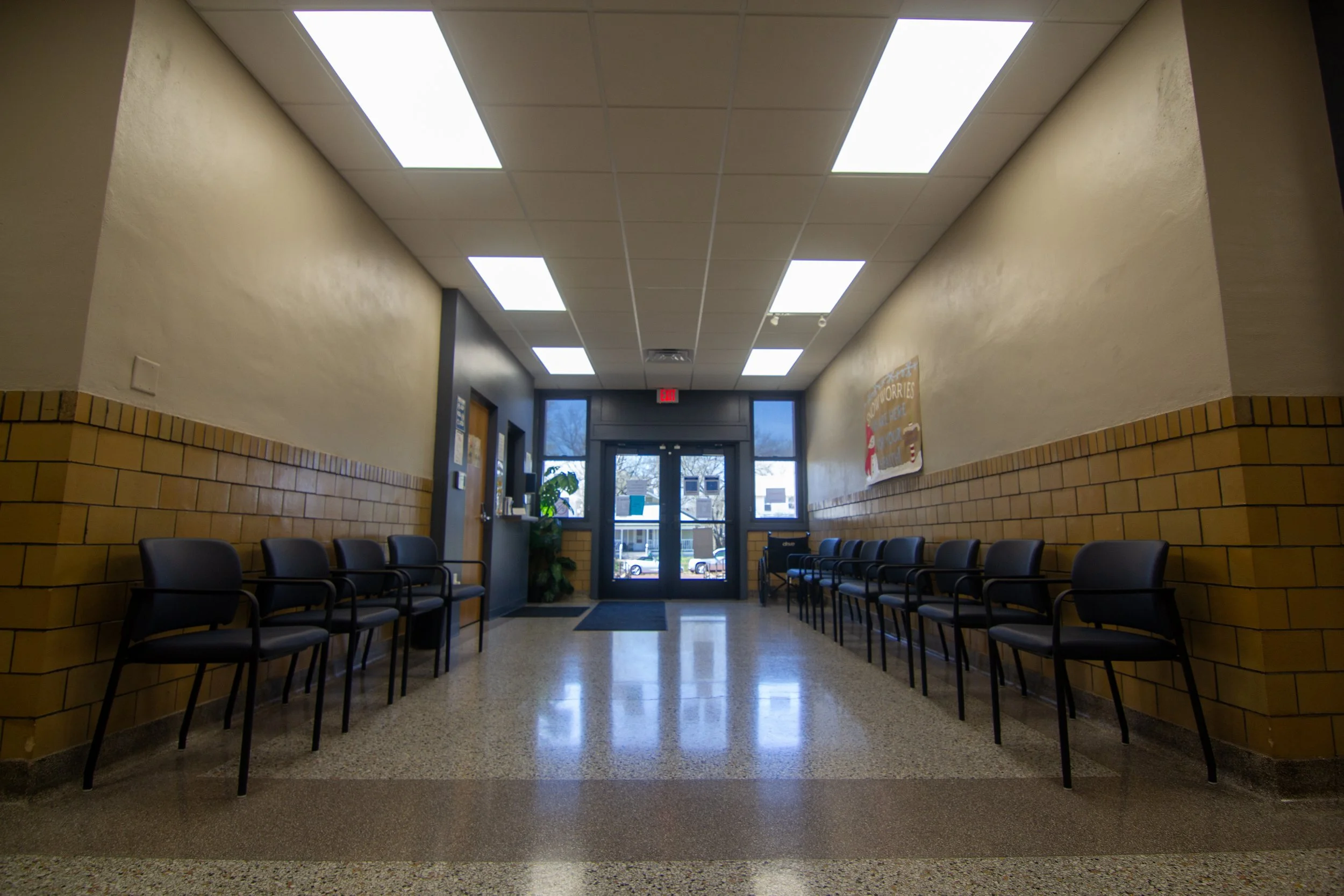 Empty waiting area with black chairs along beige and brown-tiled walls, fluorescent ceiling lights, and glass double doors leading outside, with a sign and some plants near the entrance.