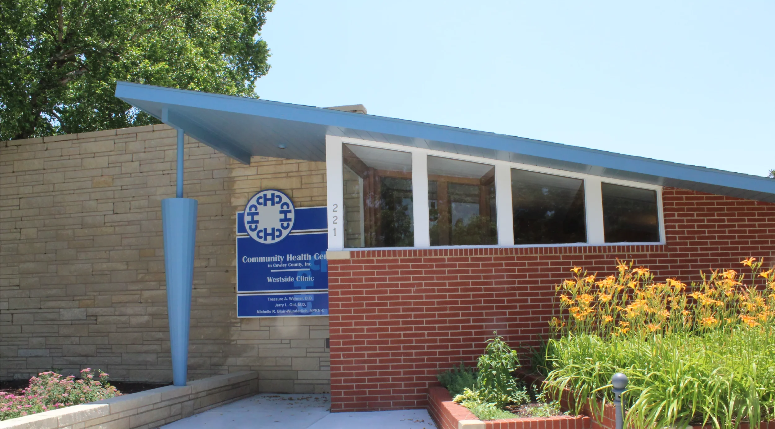 Exterior view of Community Health Center in Cowley County, with a blue sign, brick and beige wall, large windows, and yellow flowers in the garden.