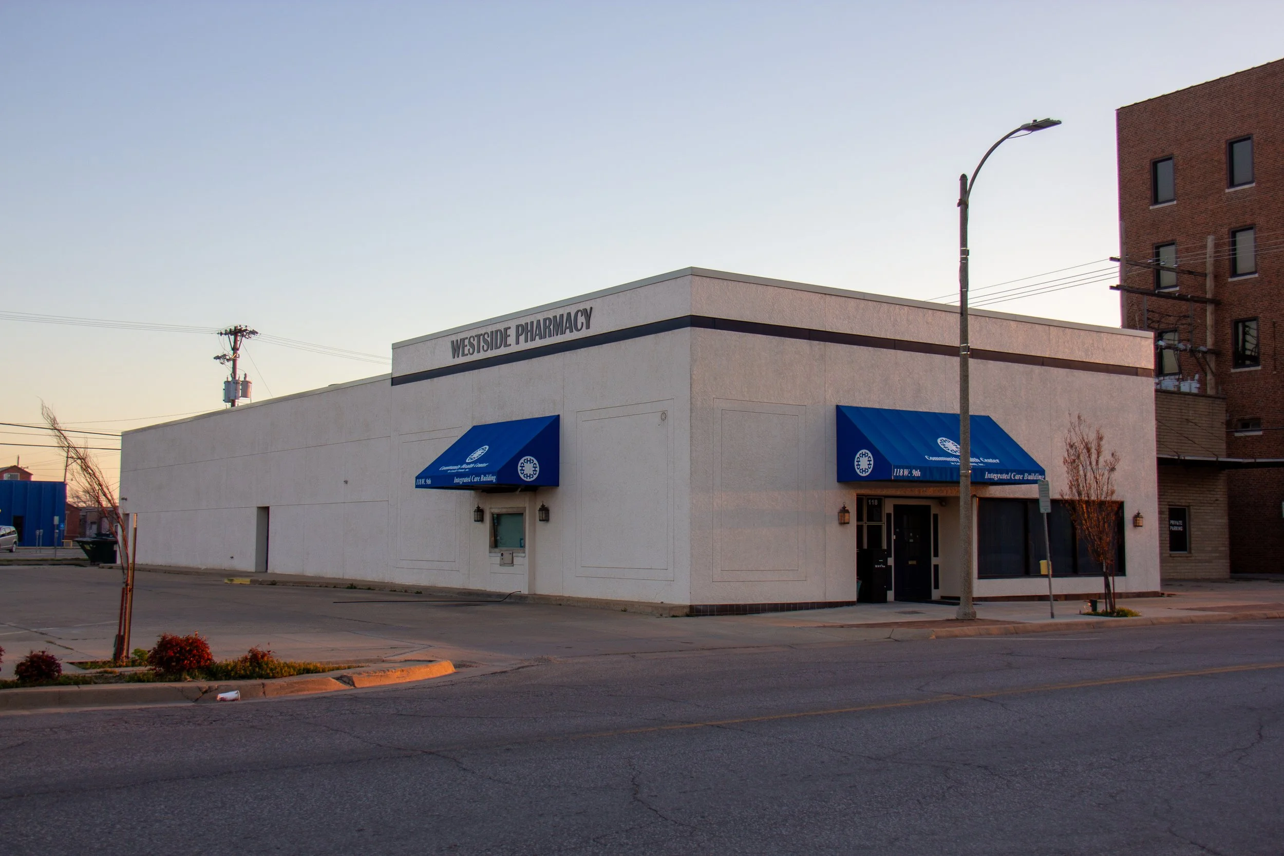 White building labeled 'Westside Pharmacy' with blue awnings, located on a quiet street with minimal traffic, under a clear sky.