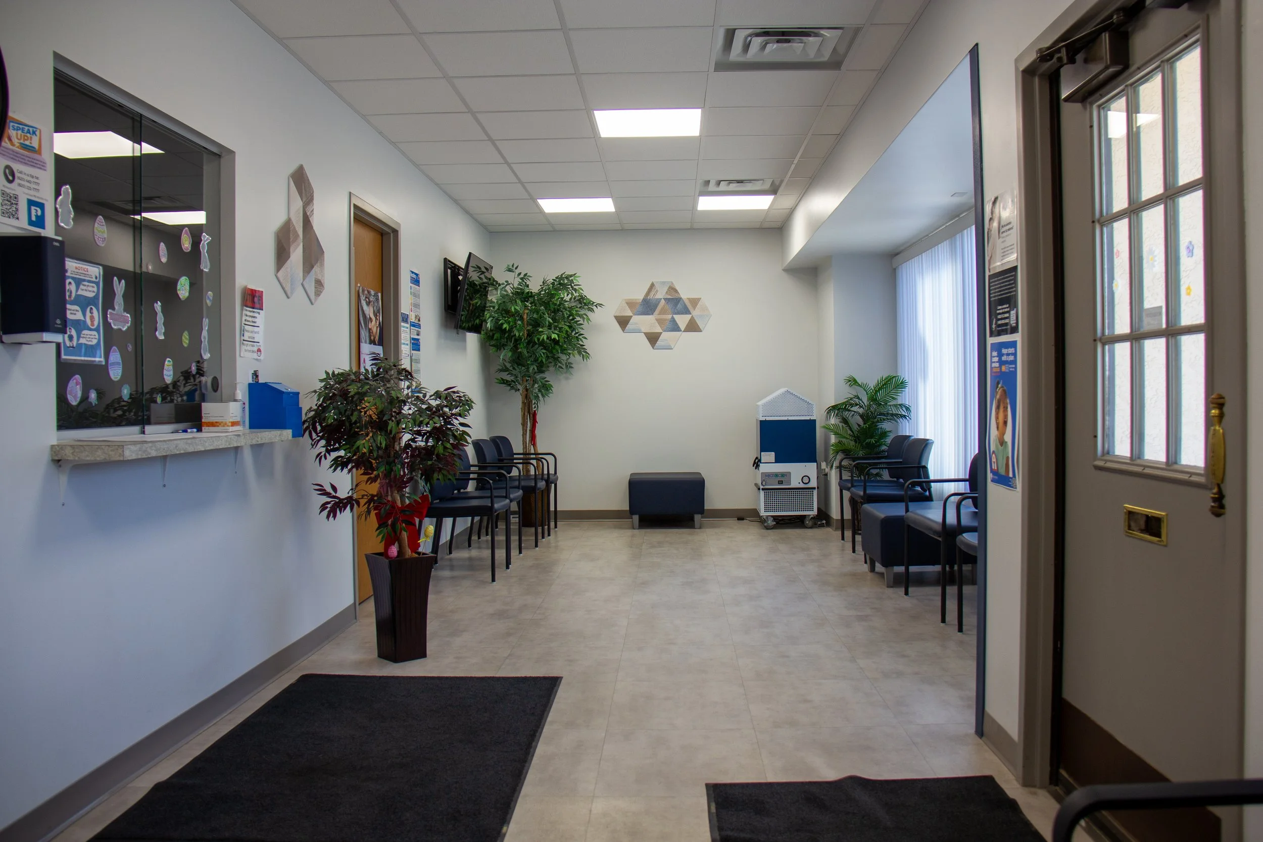 Empty waiting room with chairs, plants, wall art, and a window with vertical blinds.