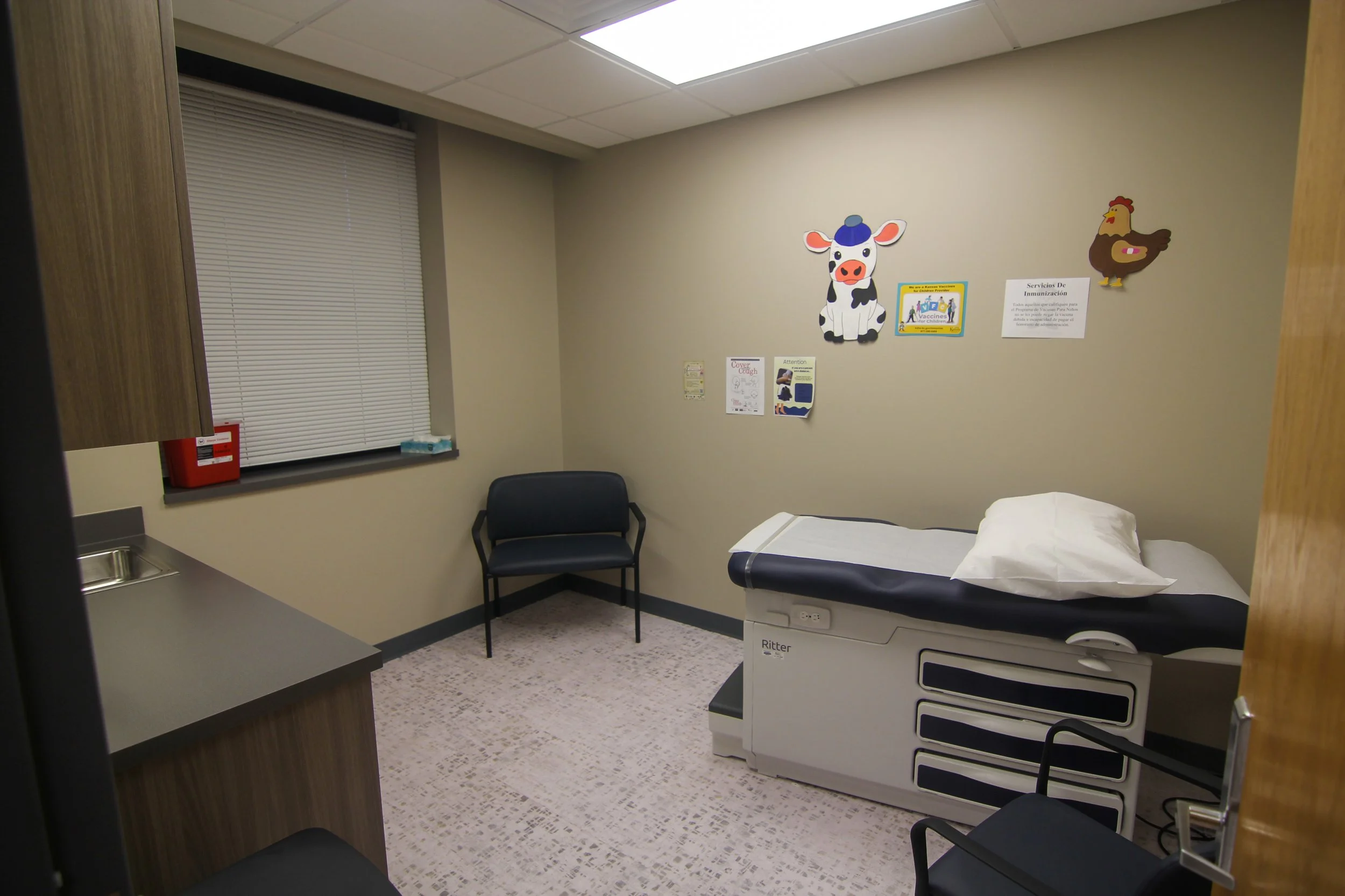 Empty medical examination room with a bed, chairs, wall decorations of a cow and chicken, window with blinds, and informational signs.