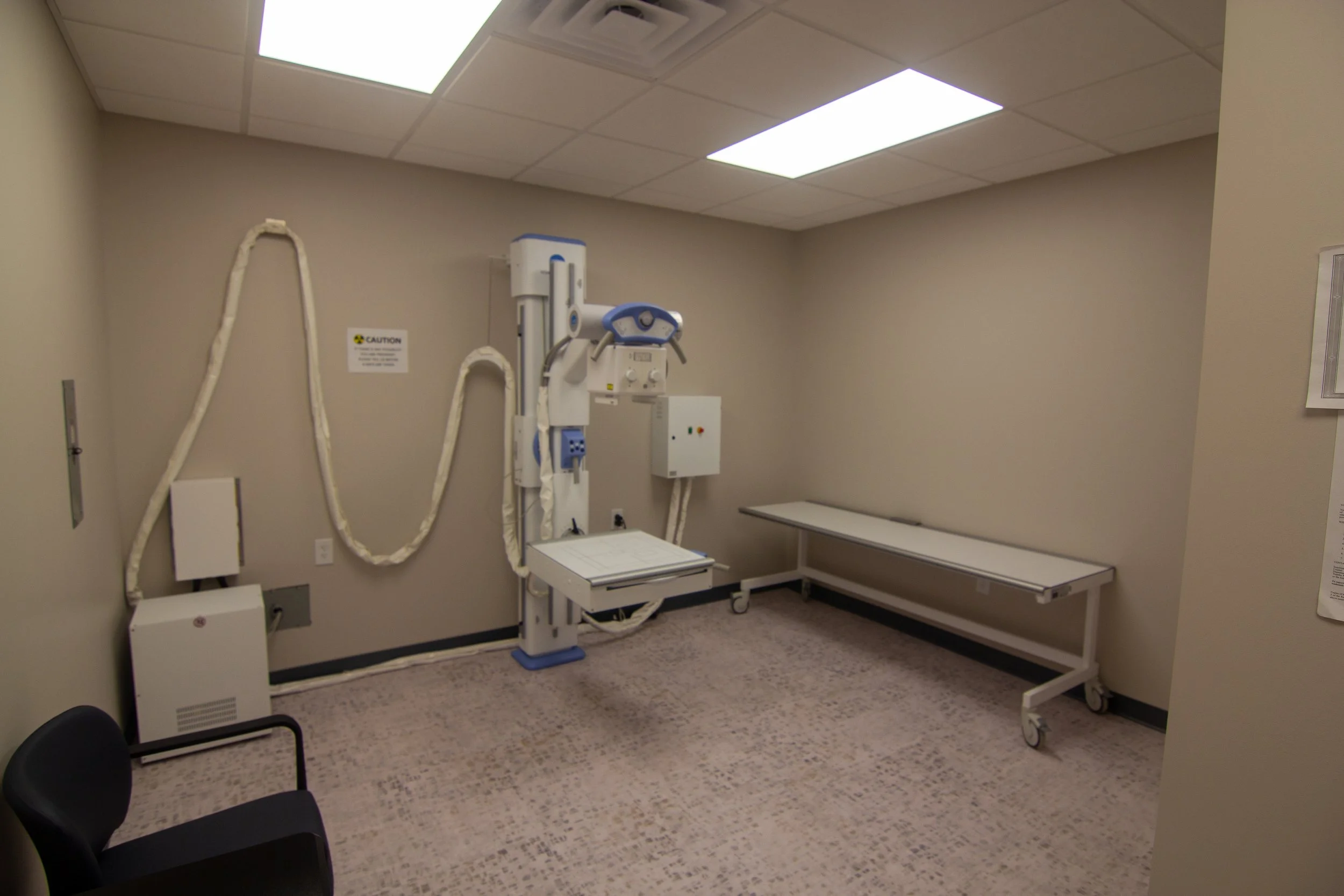 A medical imaging room with X-ray machine, patient table, and minimal furniture.