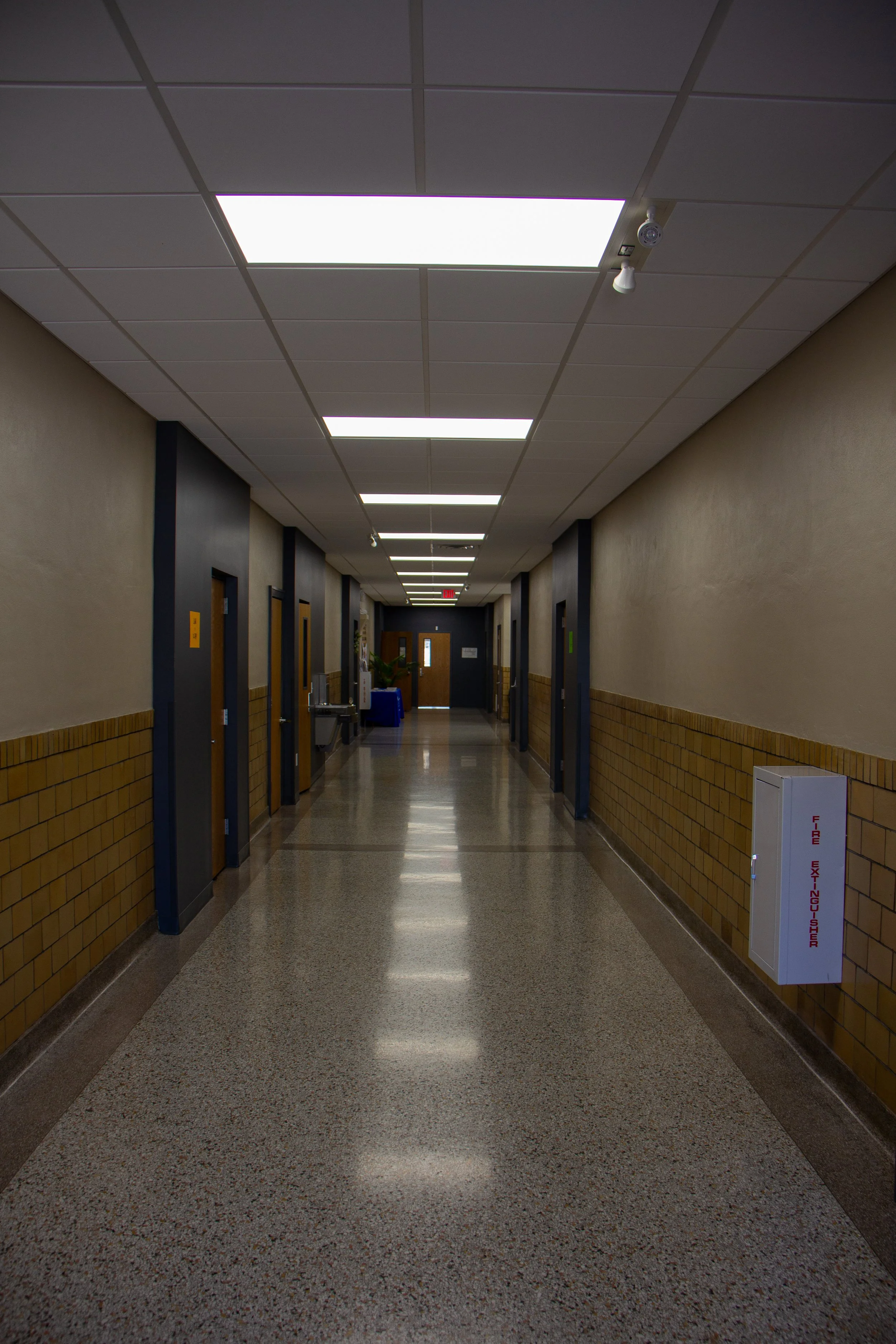 Empty hospital hallway with ceiling lights, doors on both sides, and a fire extinguisher cabinet on the right wall.