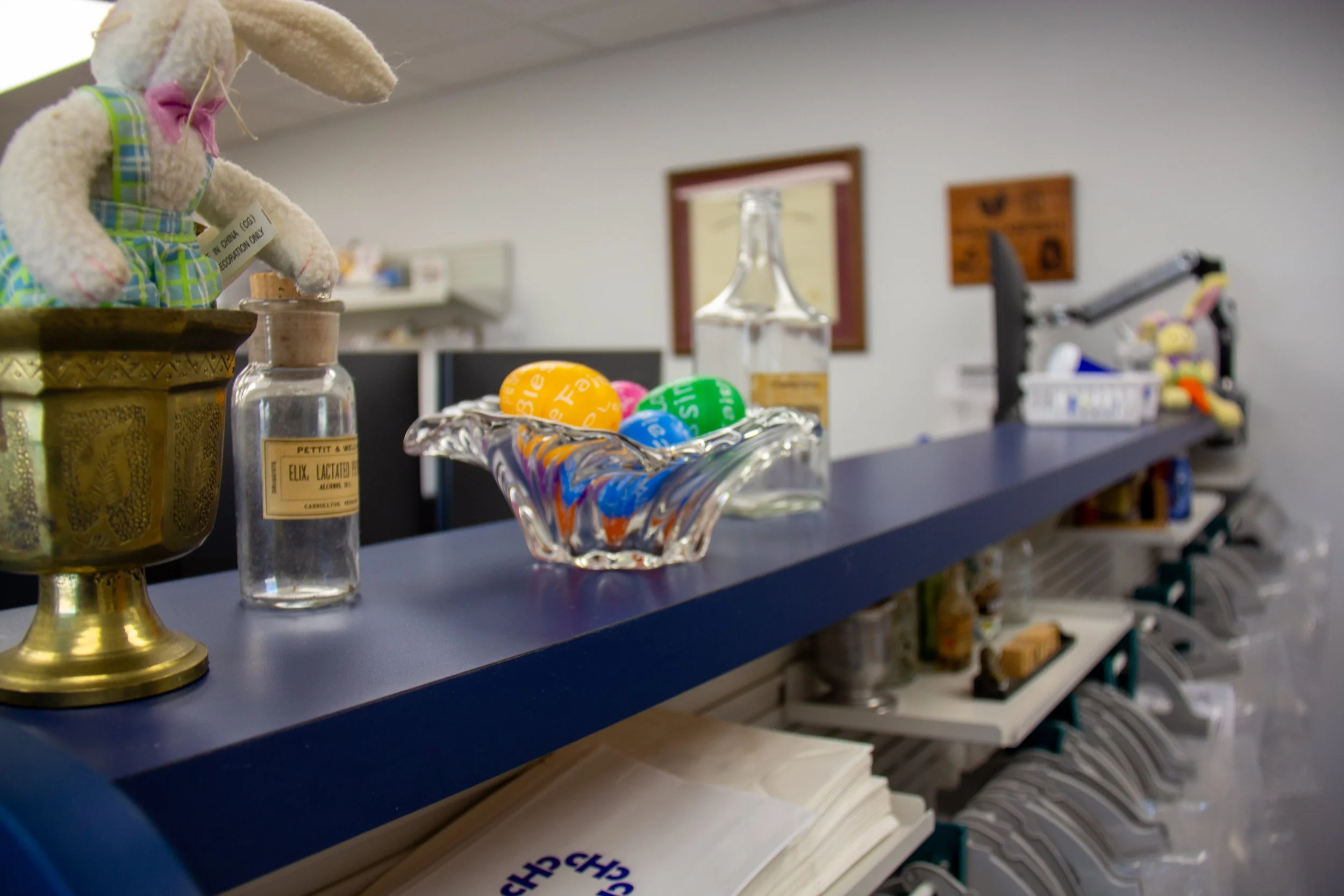 A black counter with decorative items including a plush rabbit toy, glass bottles, colorful plastic eggs in a glass dish, and medical equipment in the background, indicating a reception or workspace area.