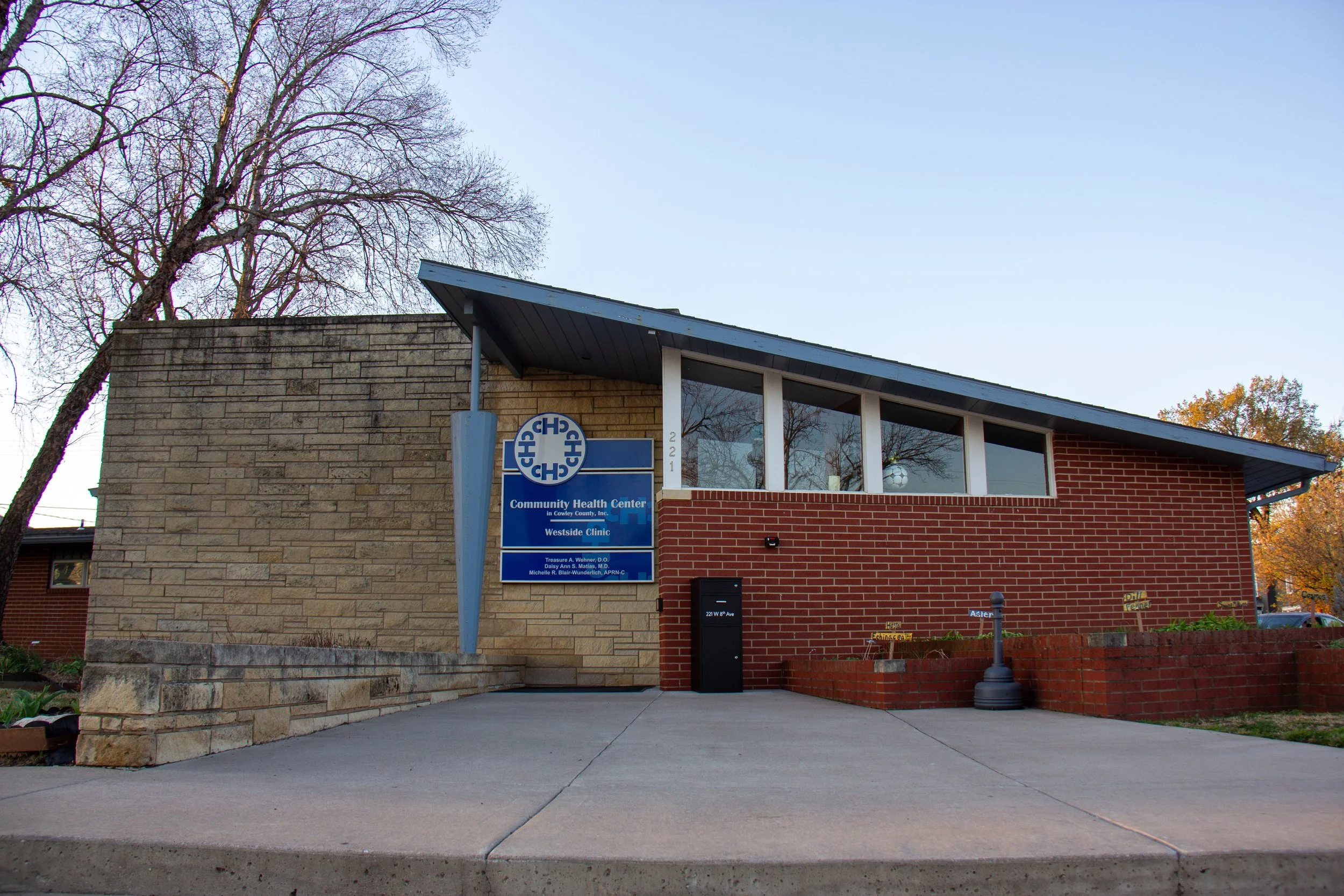 Exterior view of Community Health Center's Westside Clinic with a blue sign, brick and stone walls, large windows, and a sidewalk in front.