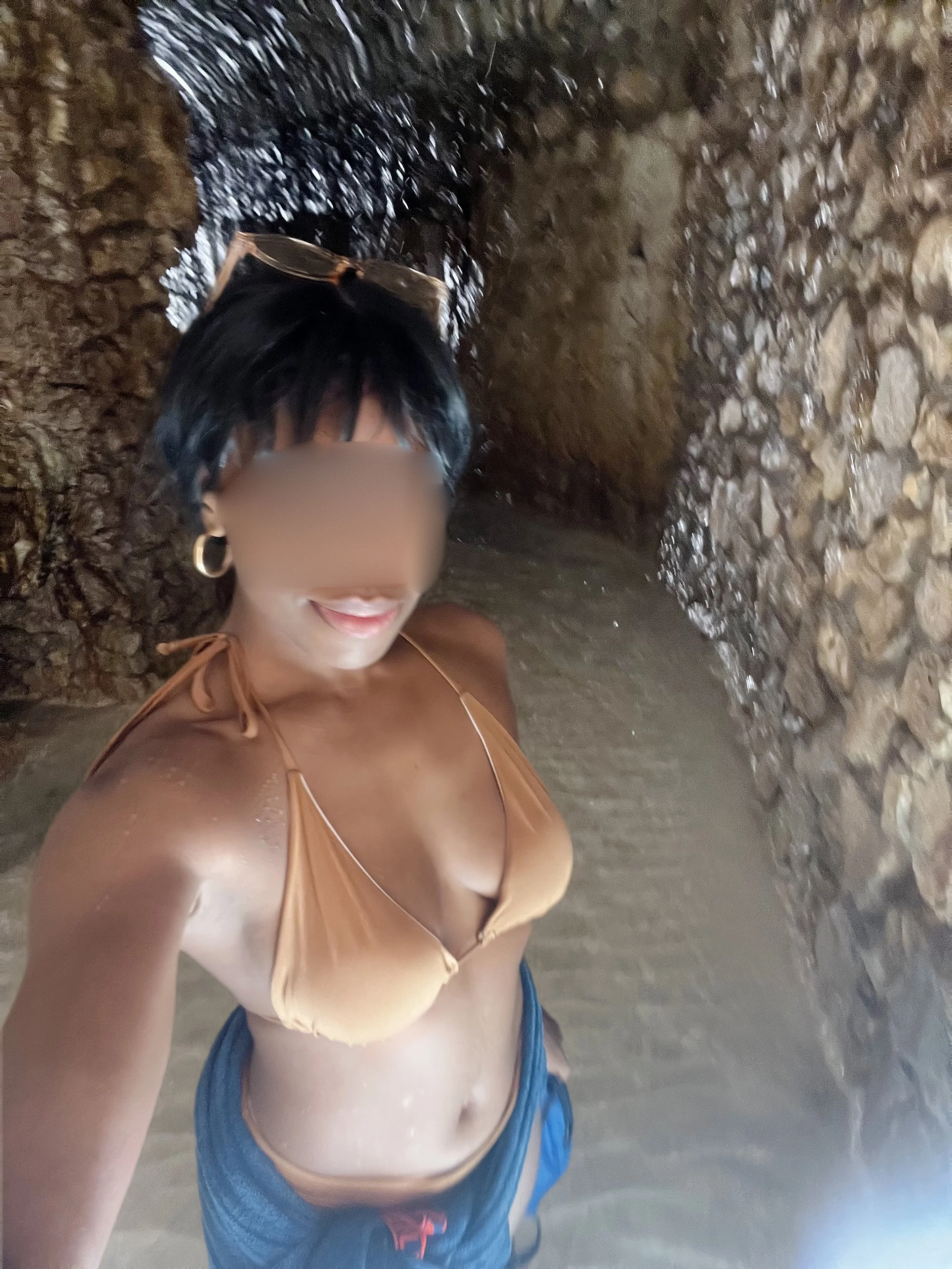 A woman with black hair and hoop earrings taking a selfie in front of a waterfall on a rocky cliffside.