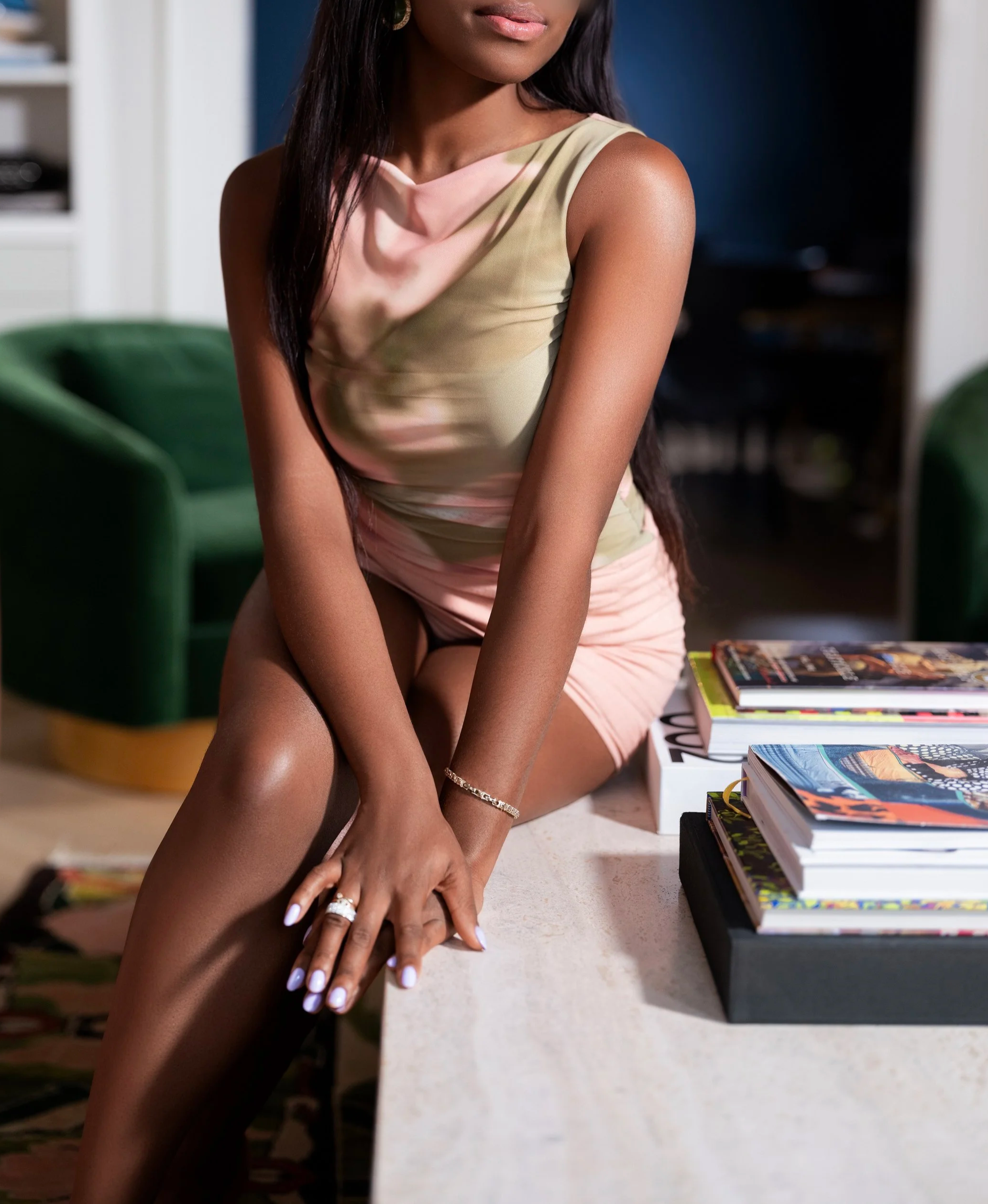 A woman with dark hair wearing a light-colored dress, leaning on a white table with her hands resting on it. There are several books stacked on the table beside her. The background features a chair and a bookshelf.