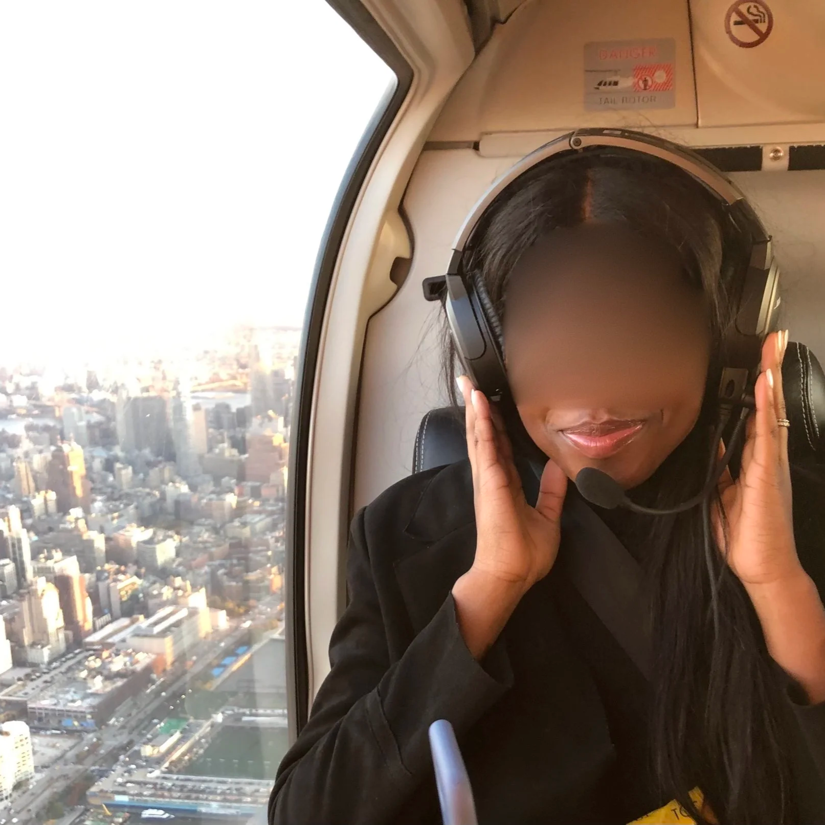 A woman with dark hair wearing headphones, sitting near a plane window with a cityscape view of skyscrapers and streets below.