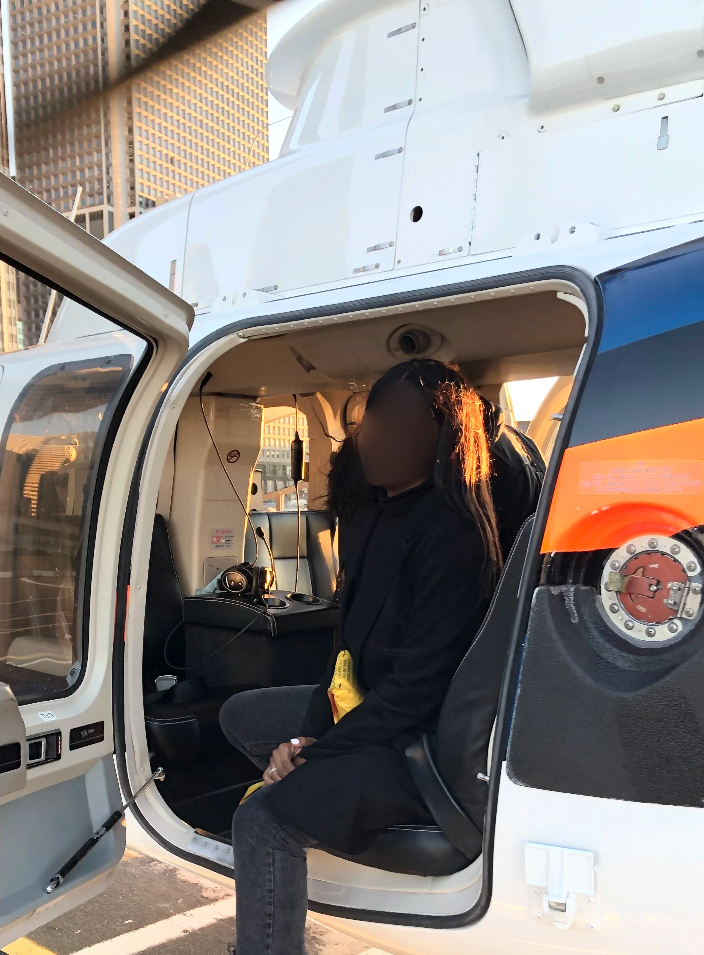 A woman with long hair, wearing a black jacket, sitting inside a helicopter with the door open, in an urban area with tall buildings in the background.