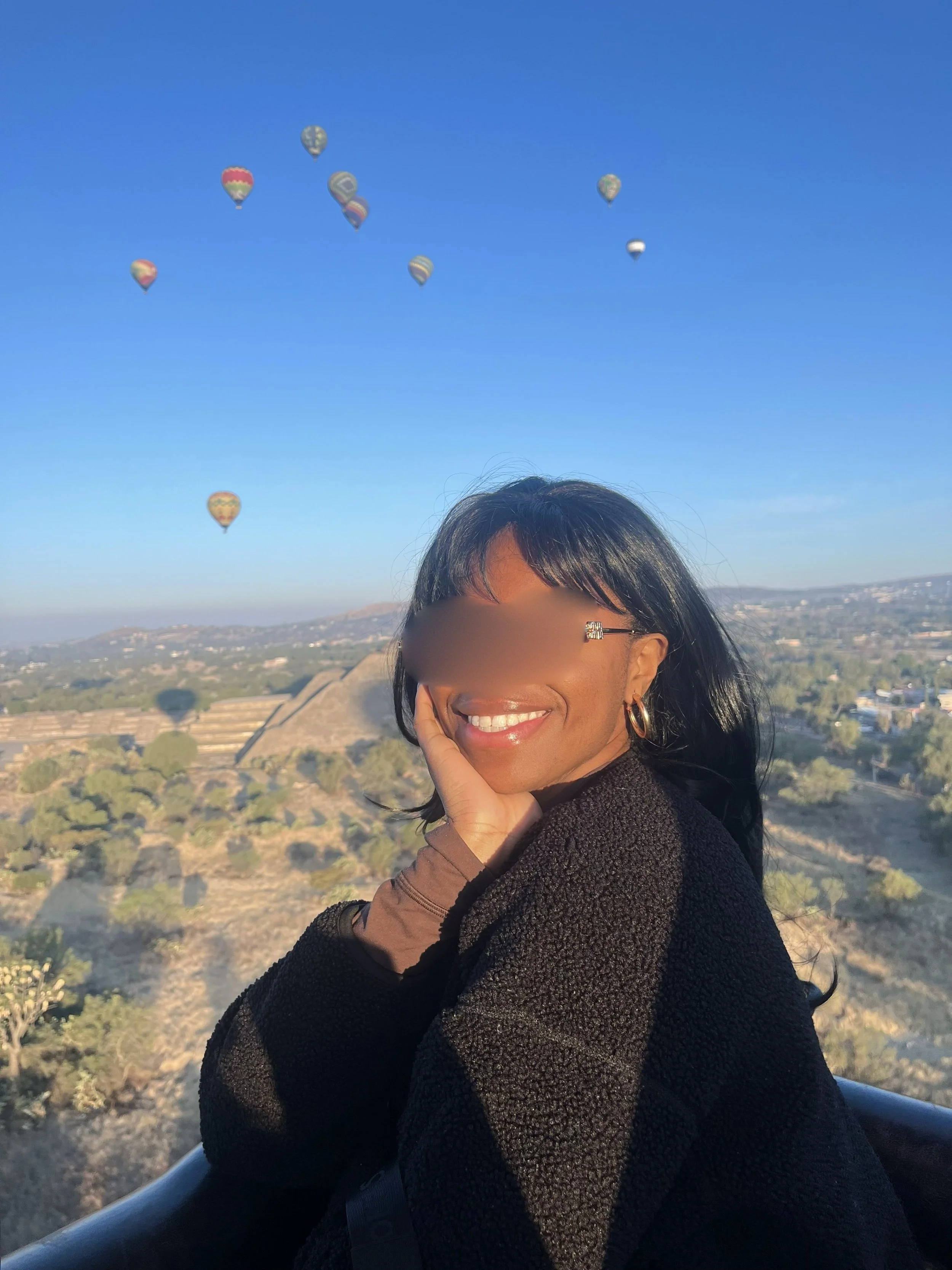 A woman smiling and resting her chin on her hand outdoors, with hot air balloons in the sky and a scenic landscape in the background.