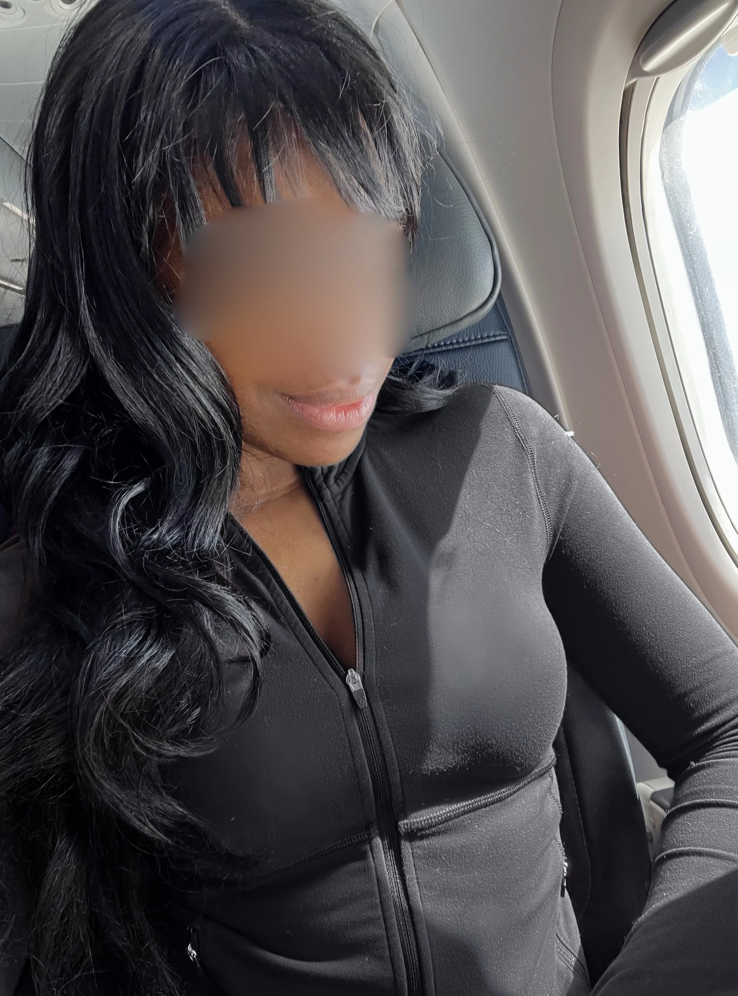 A woman with long black hair, wearing a black zip-up jacket, sitting inside a vehicle with the window partly open.