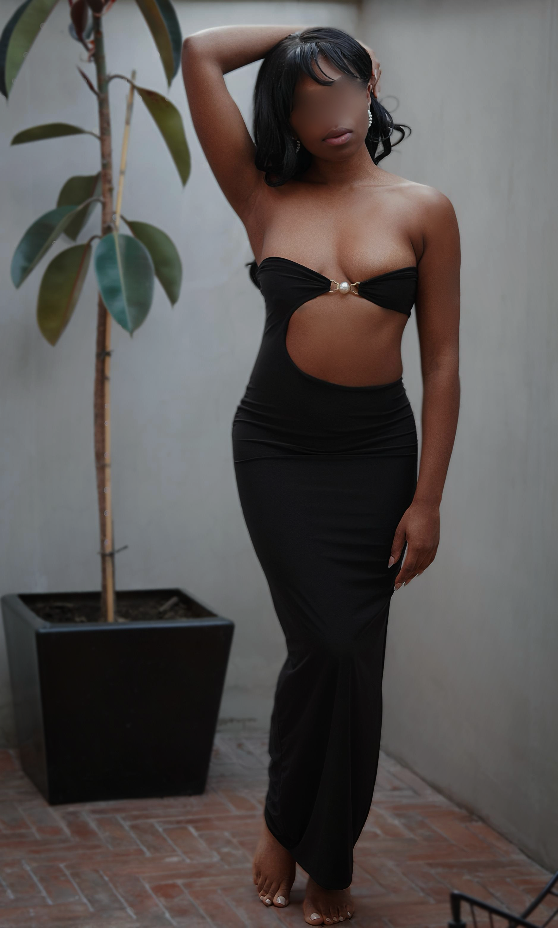 A woman with dark hair styled in waves, wearing a black strapless dress with a cutout and pearl embellishment, standing on a brick patio next to a potted plant with large, broad leaves. She has one arm raised behind her head, and her face is blurred.