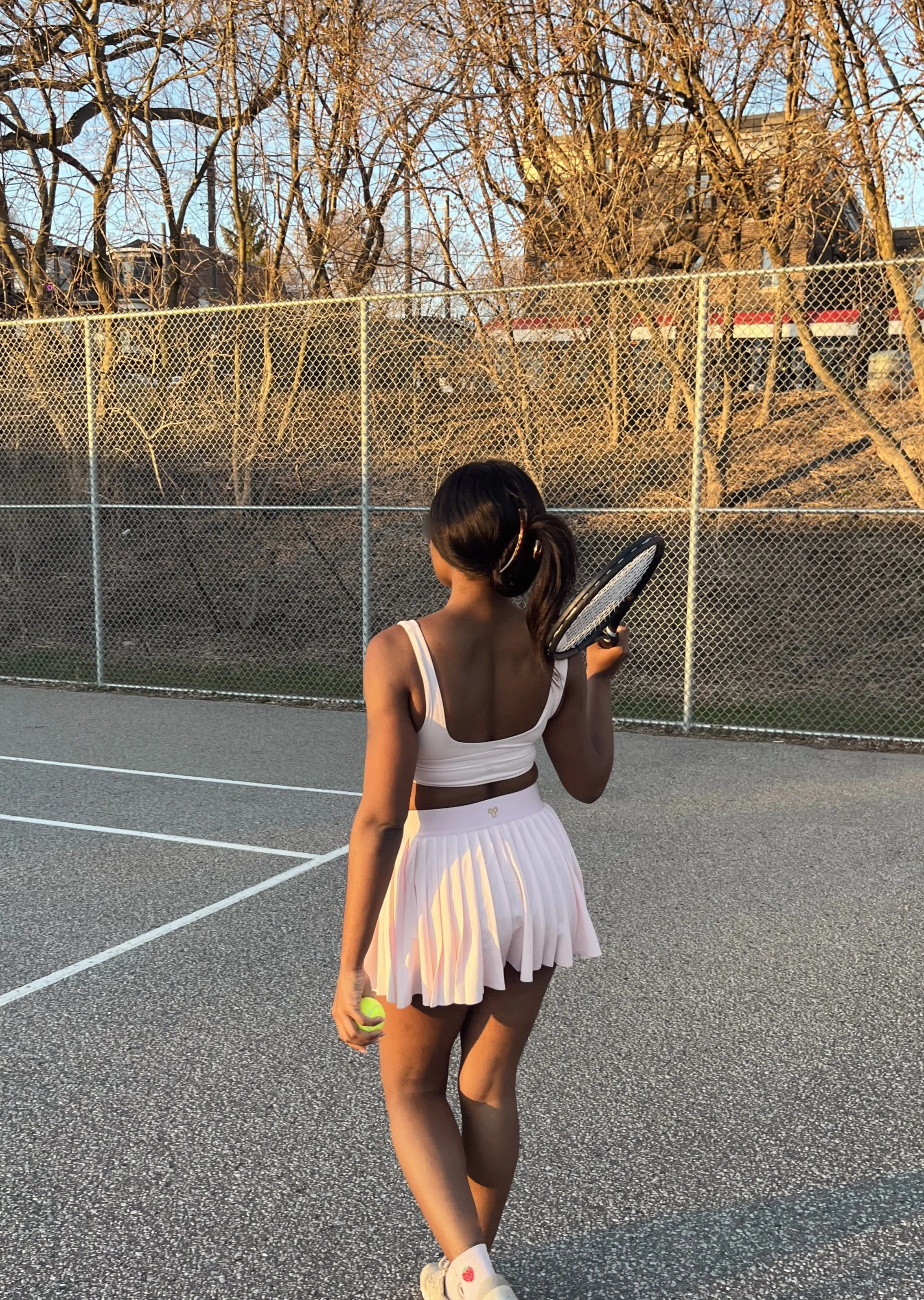 Young woman in a white tank top and pleated skirt holding a tennis ball and racket on a tennis court, with a chain-link fence and trees in the background during sunset.