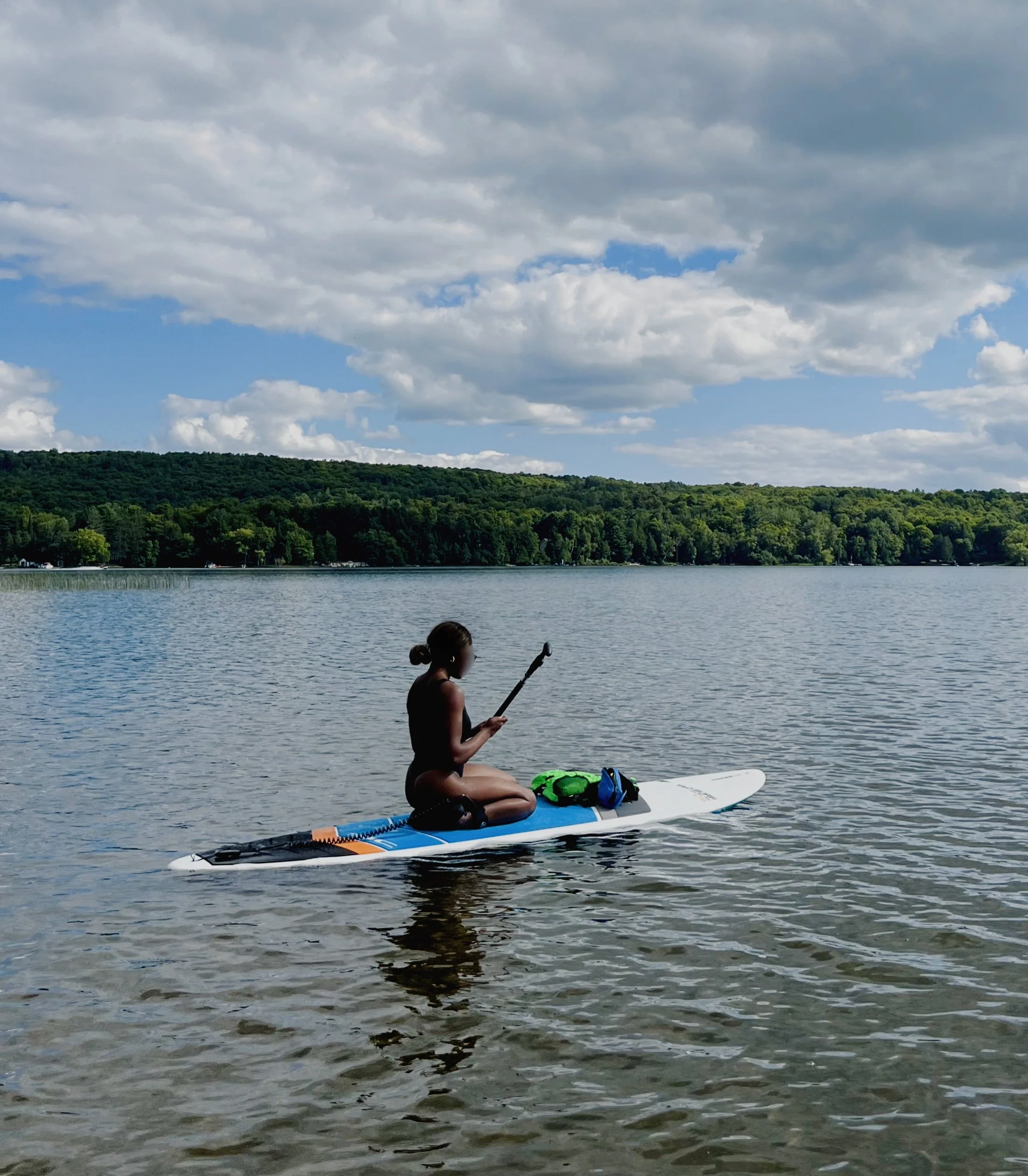 A person sitting on a paddleboard on a lake, holding a paddle, with a forest and cloudy sky in the background.