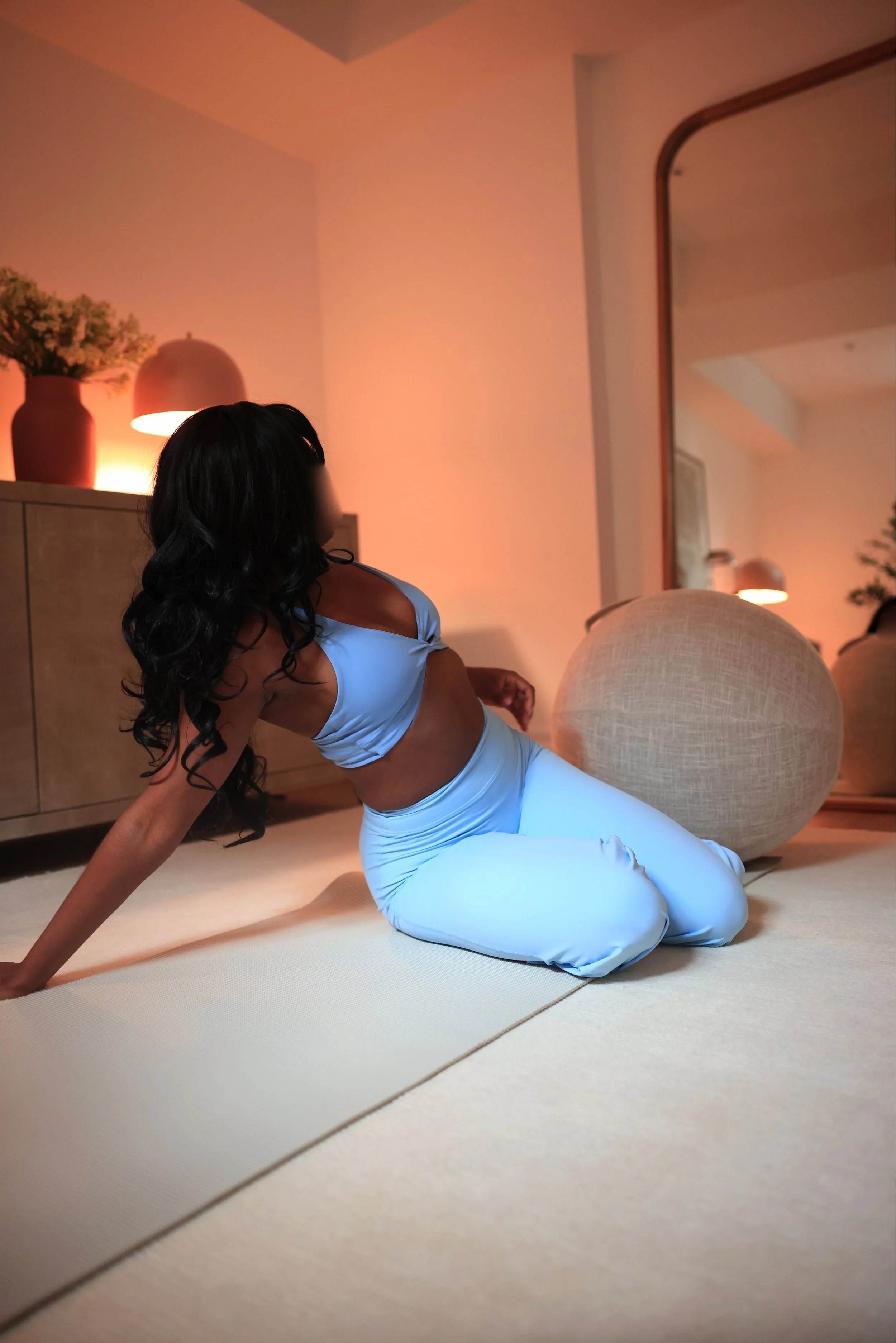 A woman with long black curly hair practicing yoga on a beige mat in a warmly lit room with modern decor. She is wearing a light blue sports bra and matching high-waisted leggings, sitting in a relaxed pose with one arm supporting her and the other resting on her leg. The background includes a wooden dresser, a vase with dried flowers, a lamp, and a mirror reflecting the room's ambiance.