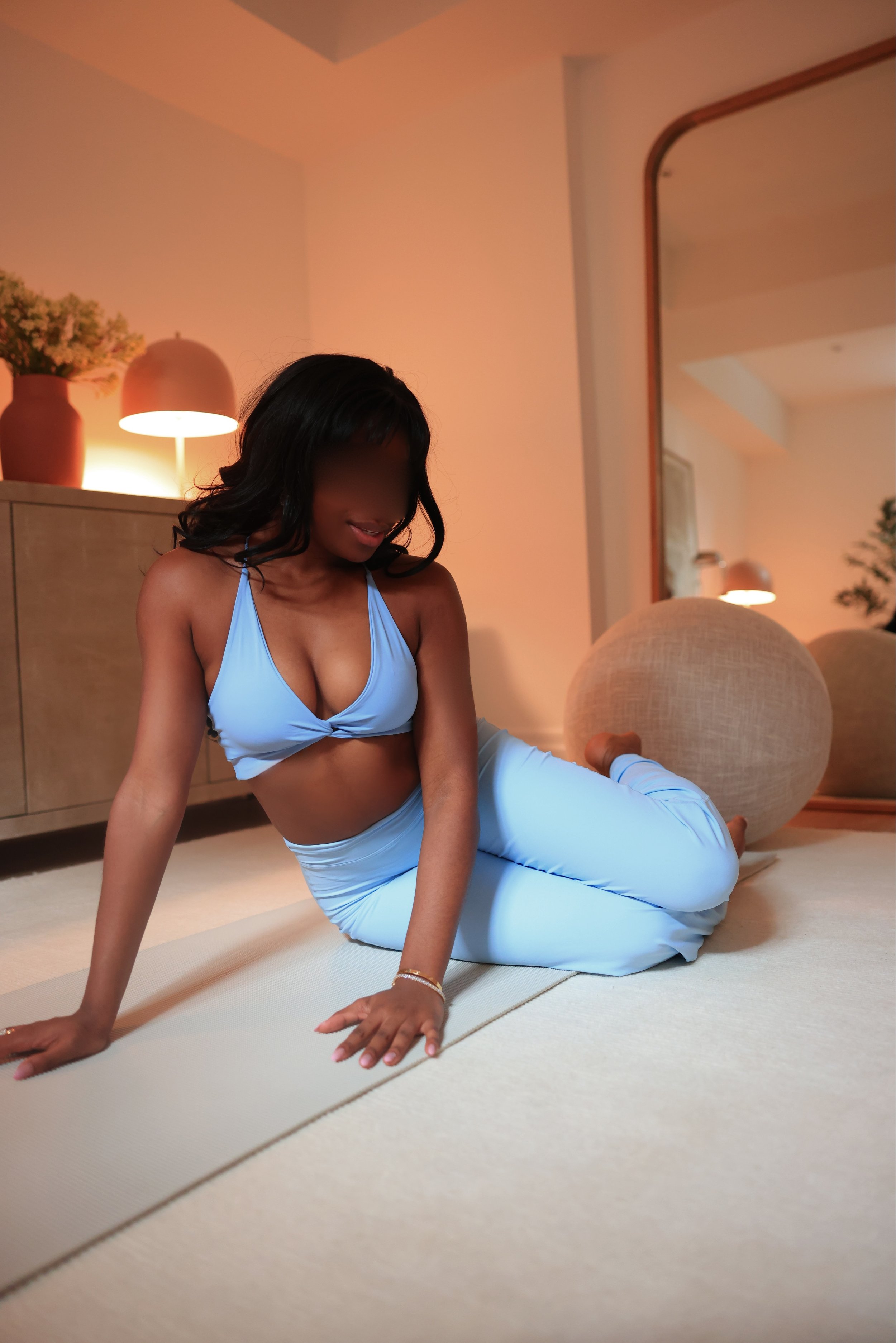 Woman in light blue workout outfit doing yoga pose on a beige mat in a cozy, well-lit room with a large mirror and decorative vases.