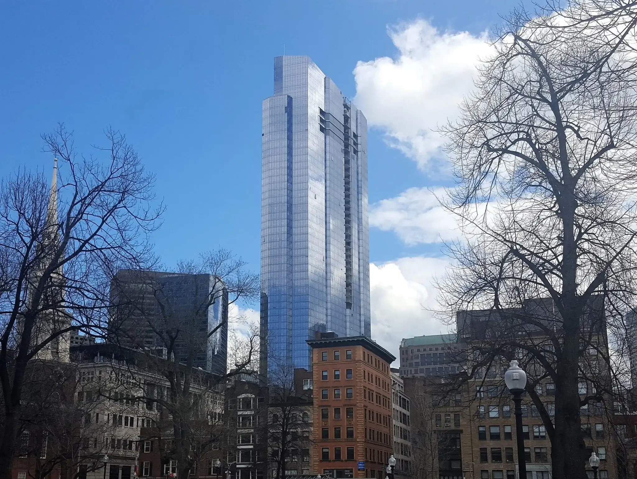 A cityscape showing a tall modern glass skyscraper surrounded by older buildings and leafless trees under a partly cloudy sky.