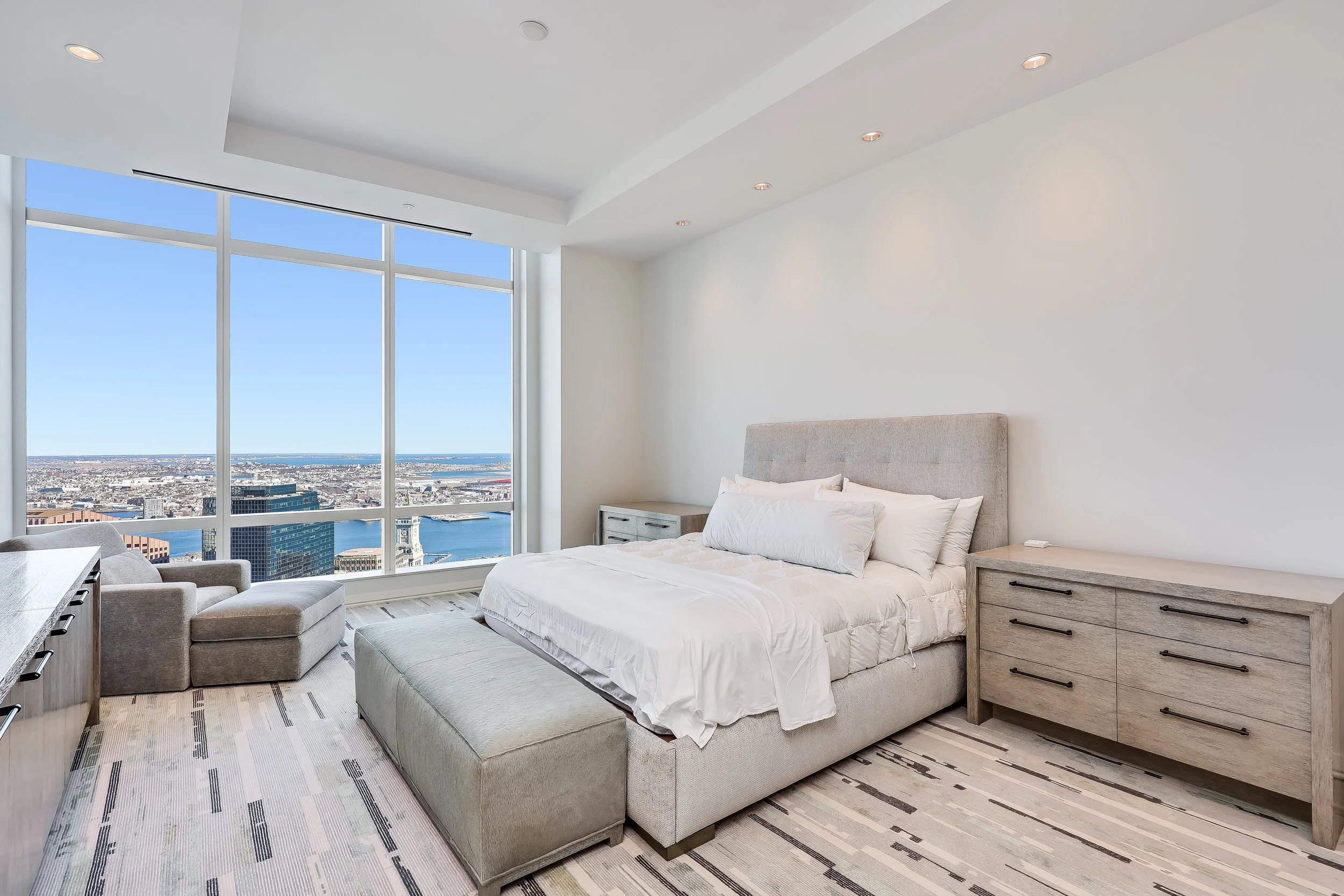 Modern bedroom with large floor-to-ceiling window overlooking cityscape and water, gray upholstered bed, white bedding, side tables, armchair, and patterned rug.