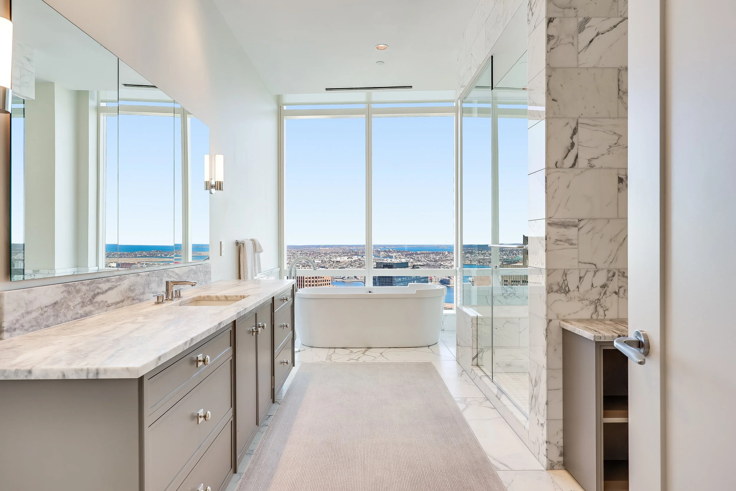 Modern bathroom with large windows showing city view, with bathtub, marble countertops, and glass shower stall.