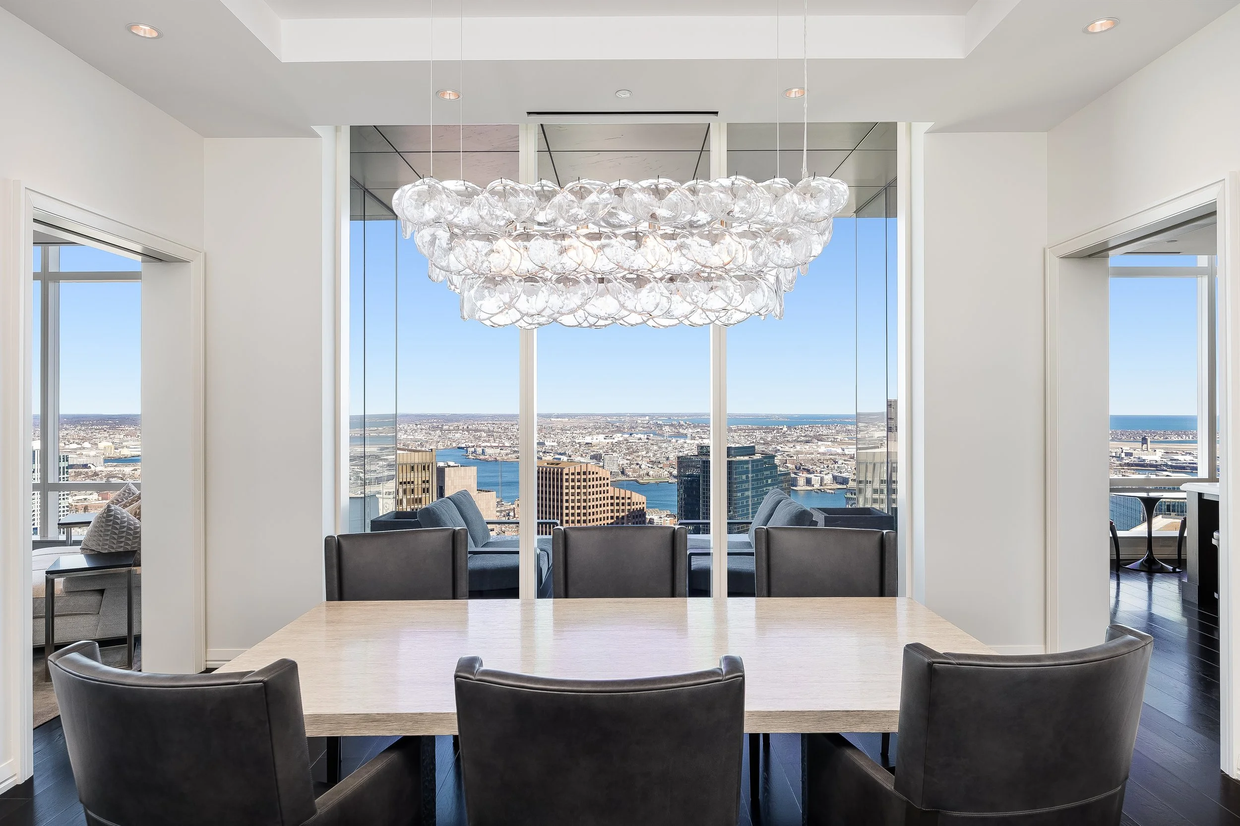Modern dining room with a large wooden table, six dark leather chairs, a chandelier made of glass globes, and floor-to-ceiling windows showing a cityscape and water view.