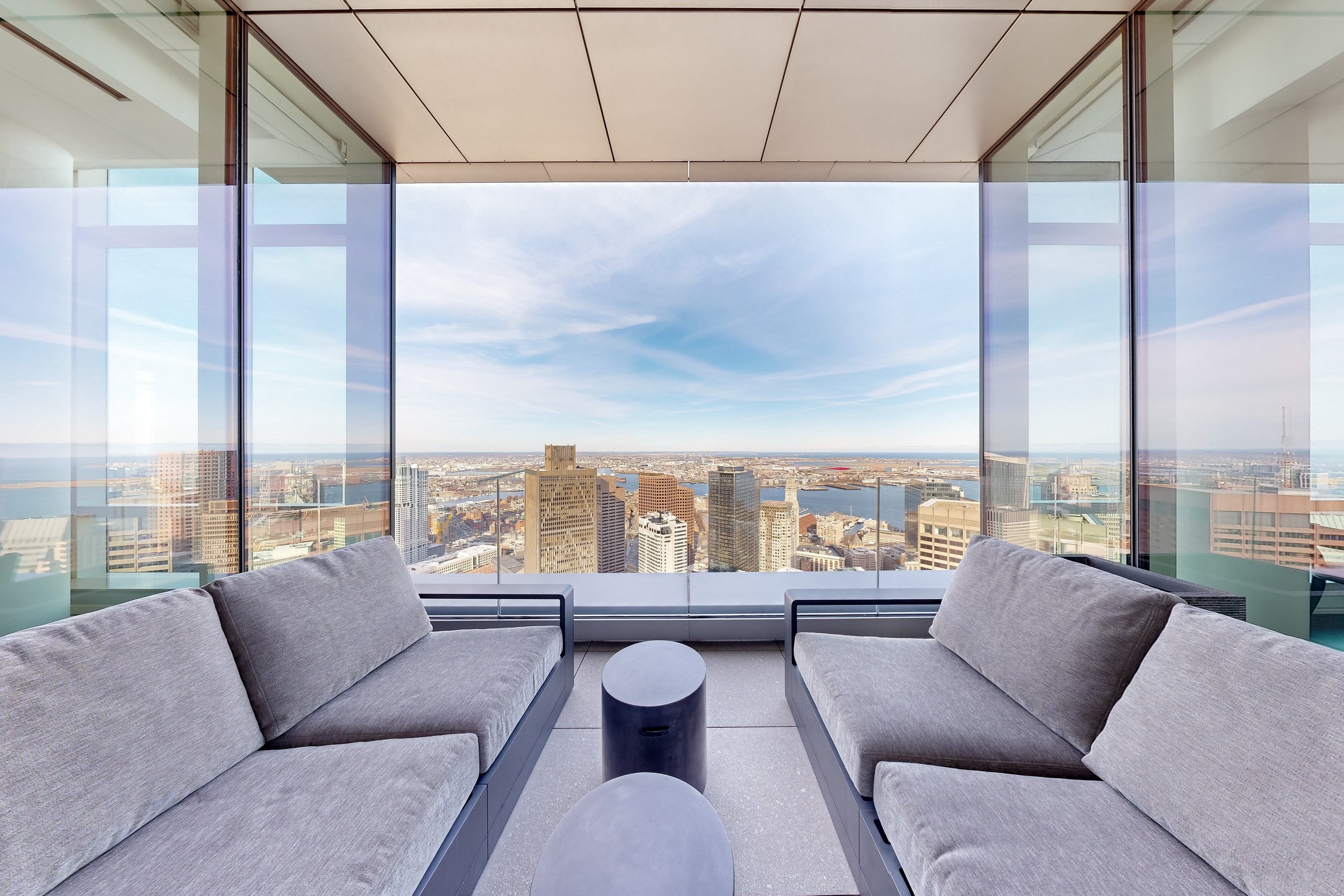 Modern lounge with two gray sofas facing each other and a small round table in the middle, overlooking a city skyline and water through floor-to-ceiling glass windows.