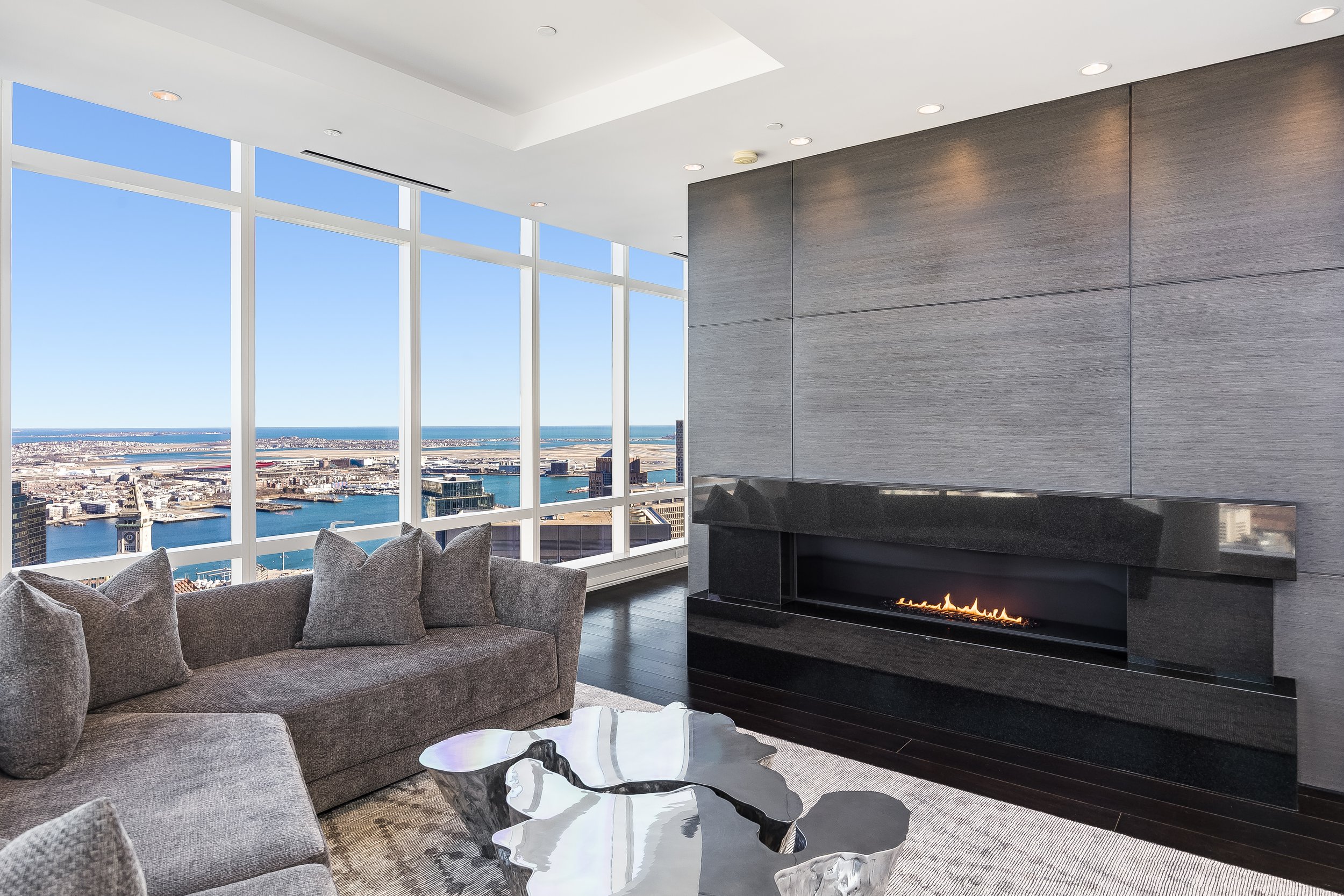 Modern living room with large windows overlooking a city skyline and water, with a gray sofa, marble coffee table, and a sleek fireplace.