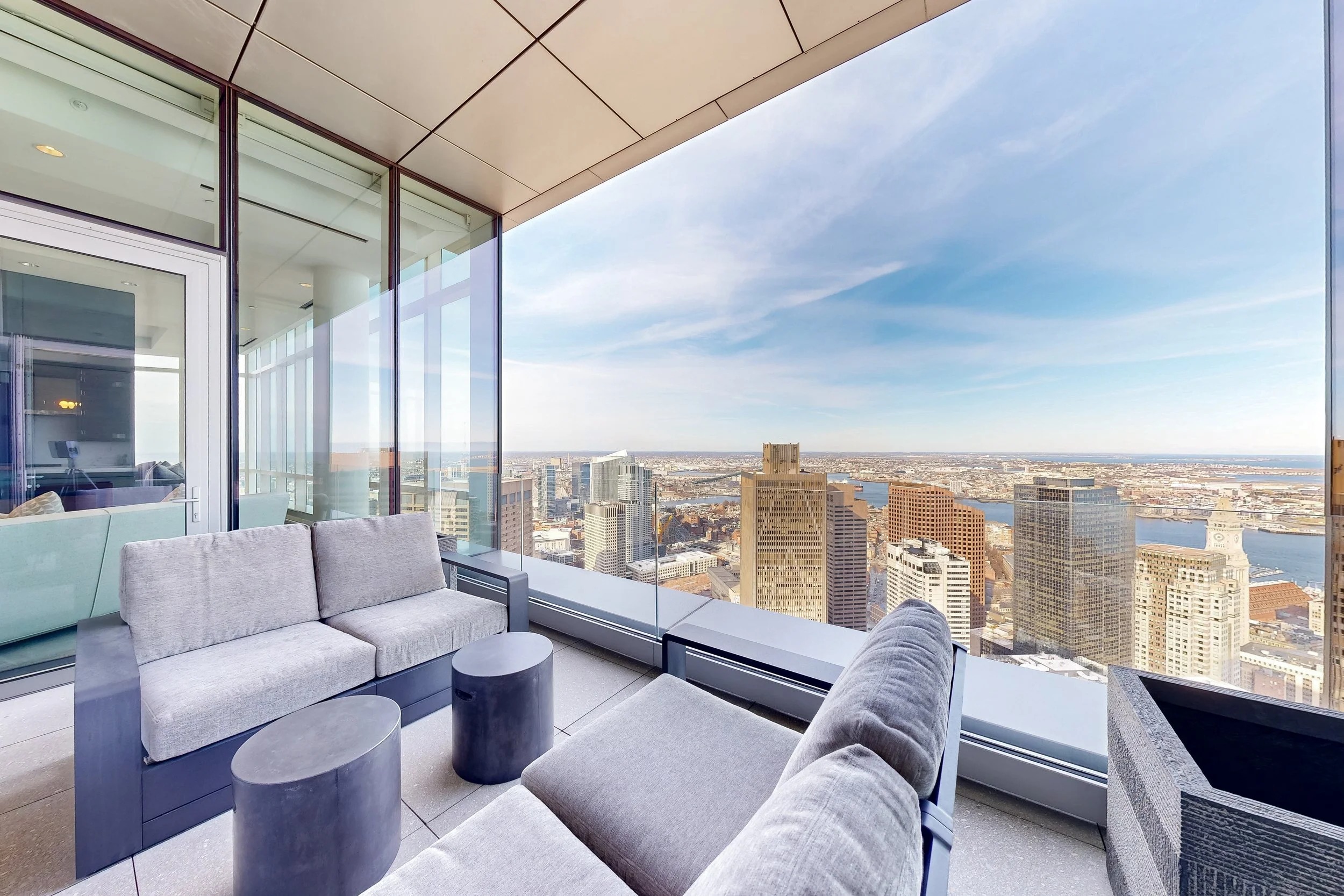 Balcony with gray cushioned sofa and tables overlooking a city skyline with tall buildings and a river with blue sky and few clouds.