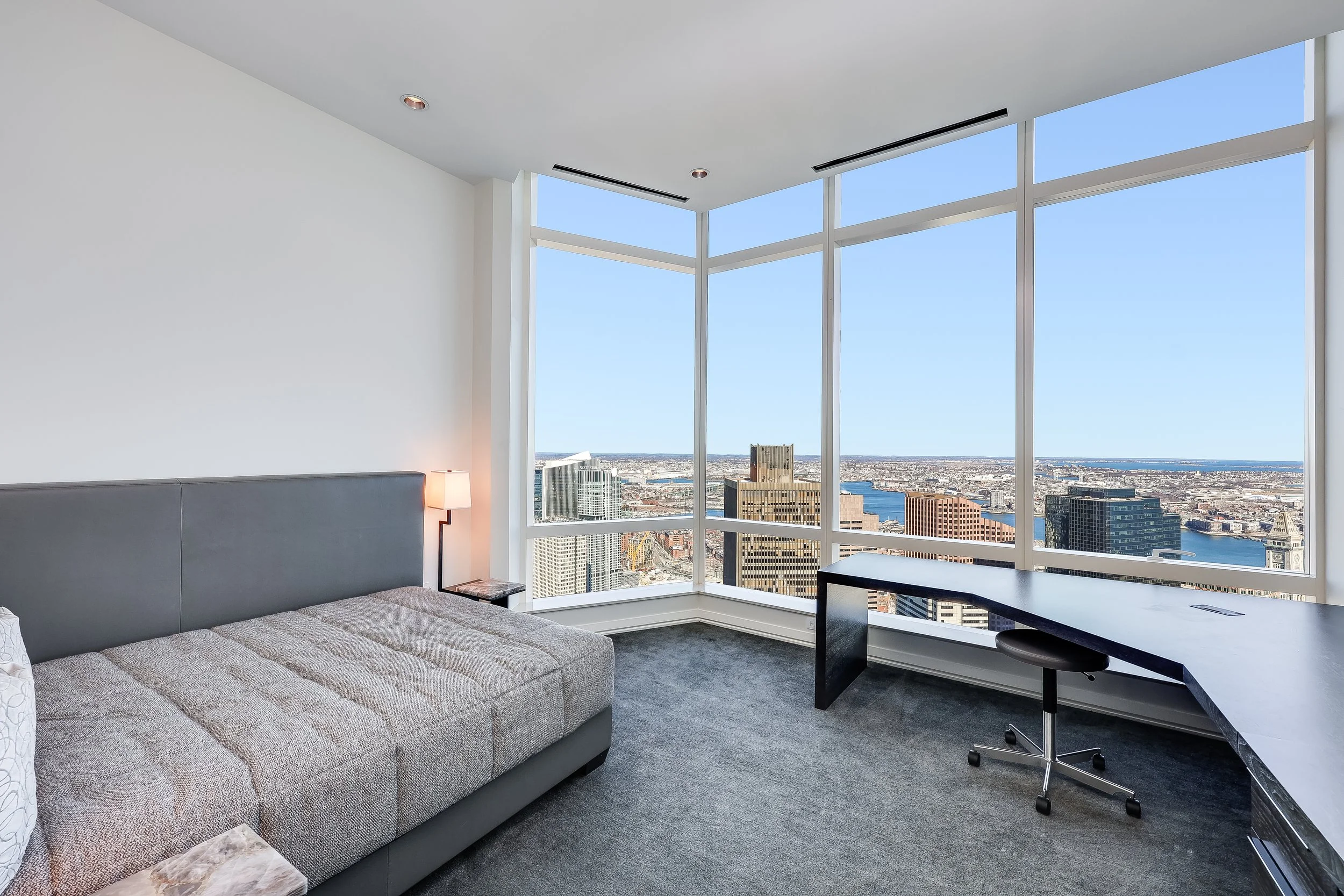 Modern high-rise apartment with a view of the city skyline and river through large corner windows, featuring a gray sofa, a small side table with a lamp, and a black curved desk with a rolling chair.