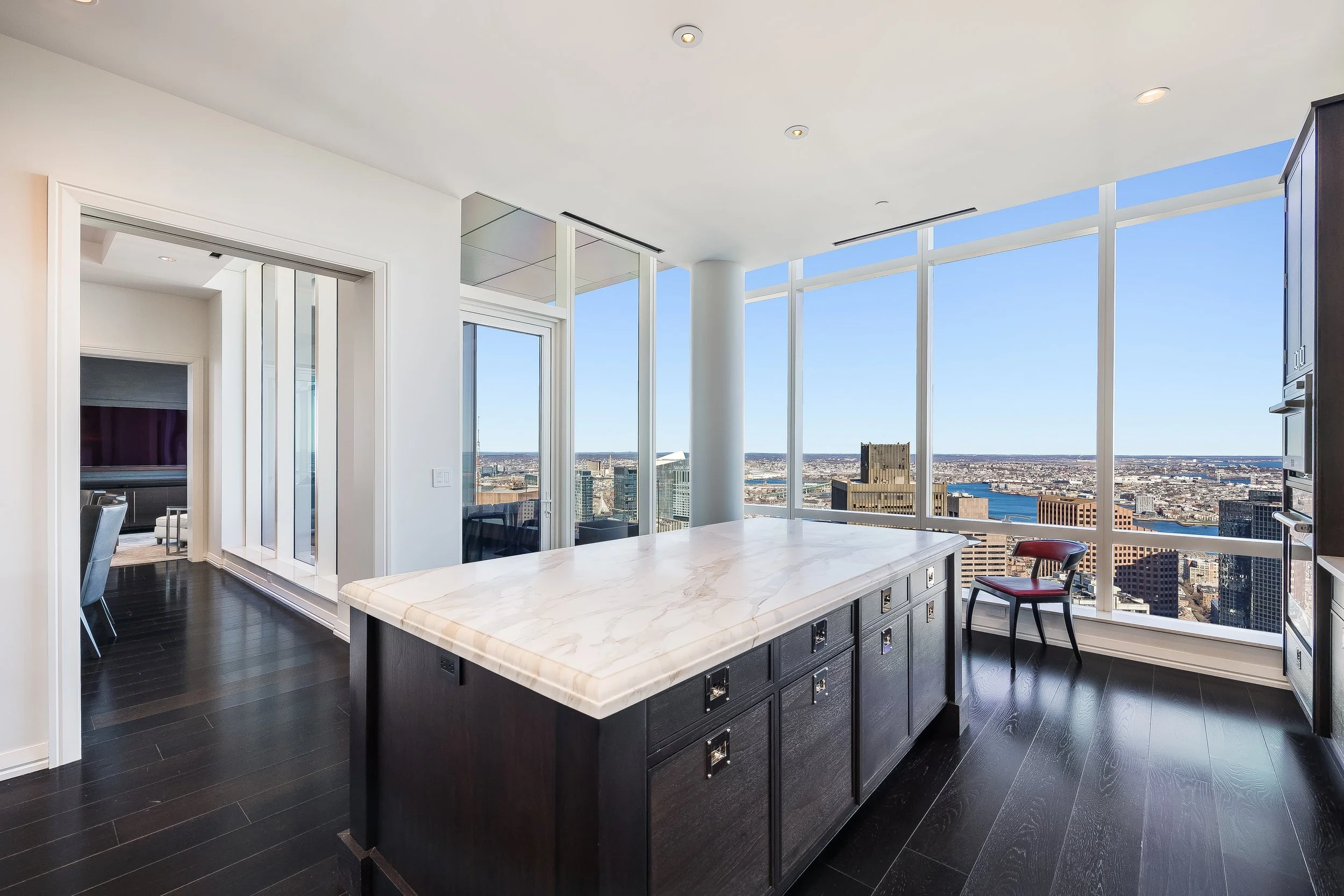 Modern kitchen with large windows and cityscape view, black kitchen island with marble top, dark wood floors, and a single red chair.