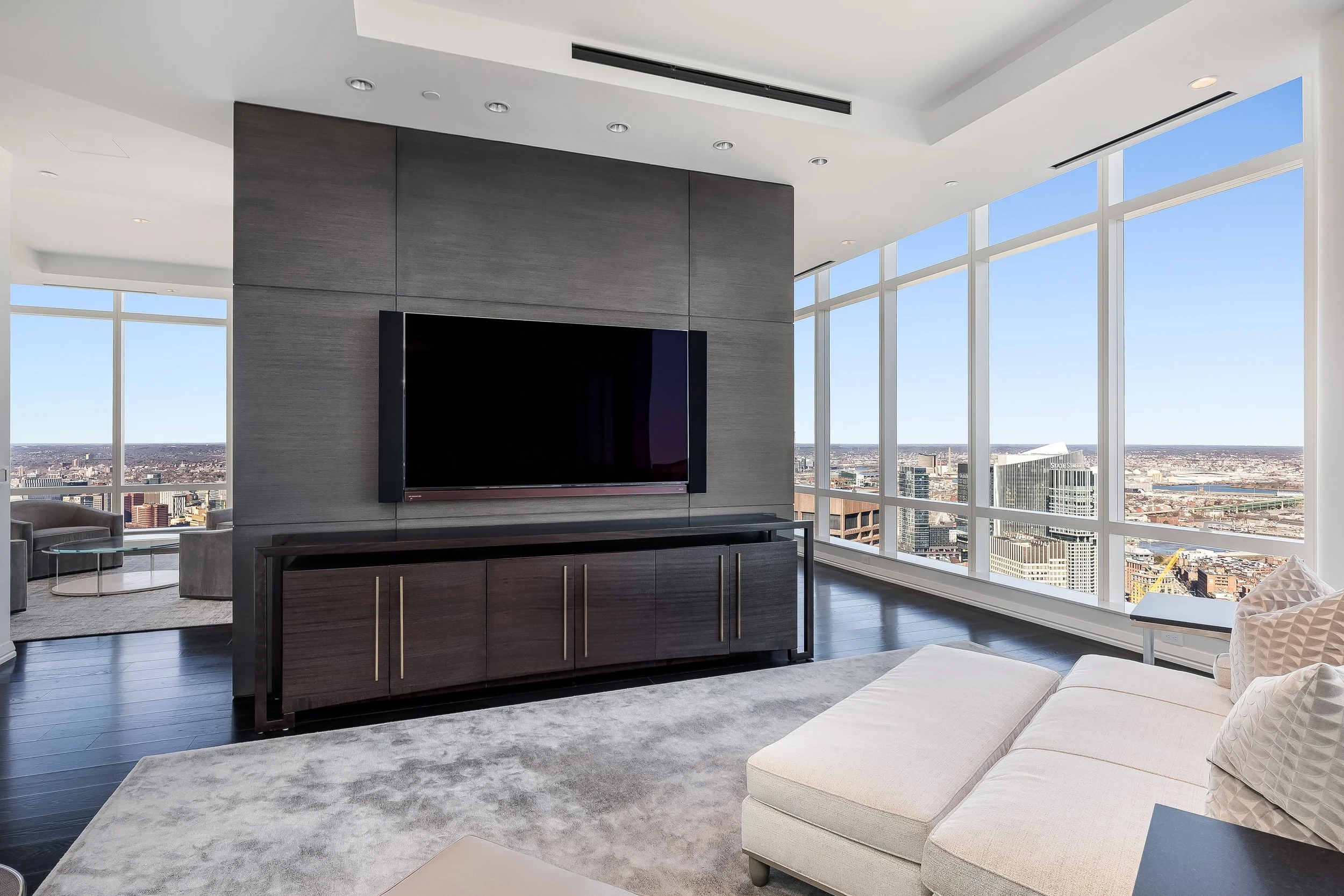 Modern living room with large floor-to-ceiling windows offering city skyline views, a mounted flat-screen TV on a wooden wall, and neutral-colored furniture.