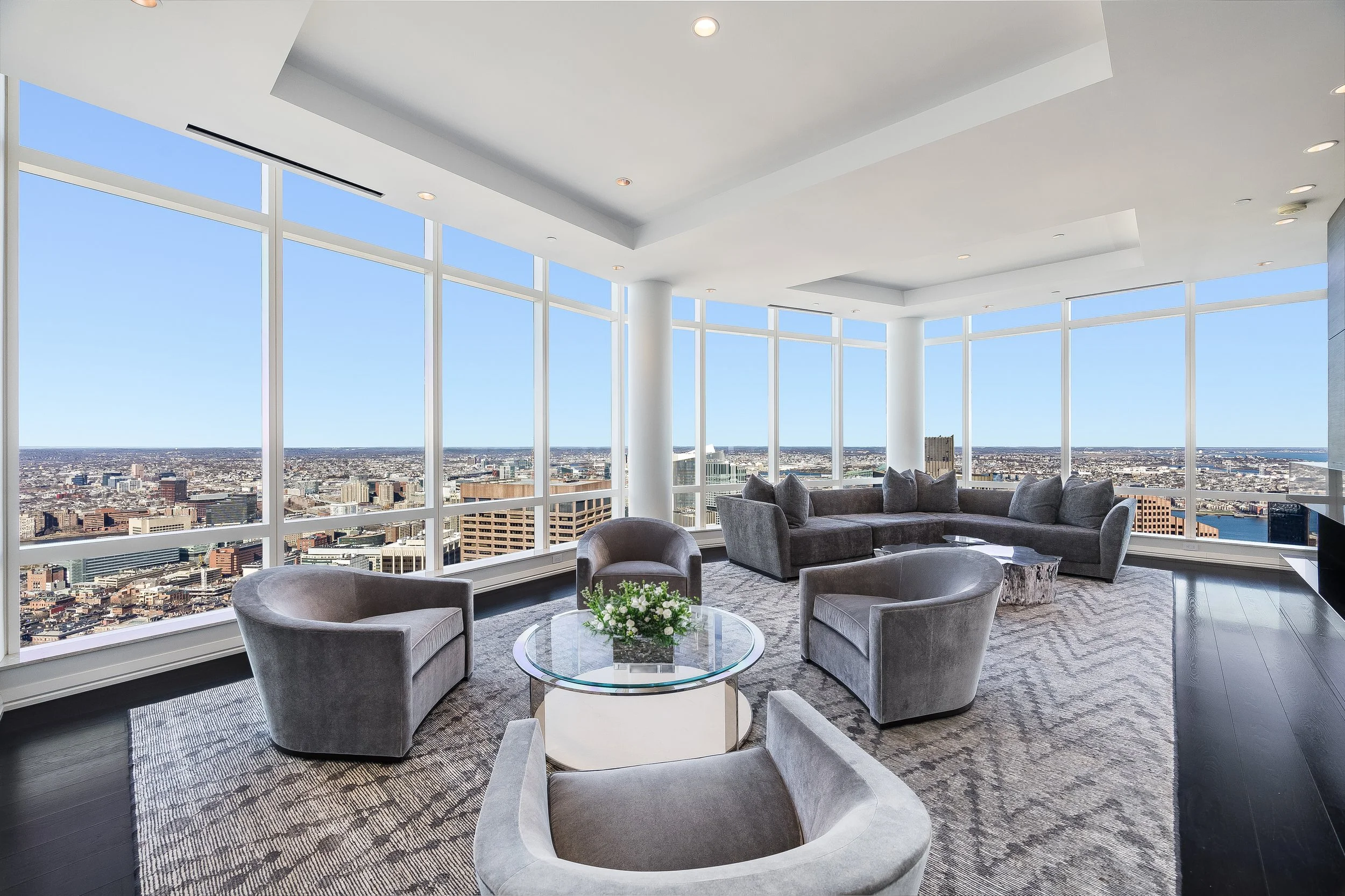Luxury high-rise lounge with floor-to-ceiling windows overlooking a city skyline, furnished with gray velvet chairs, a sectional sofa, a glass coffee table with a floral arrangement, and a patterned rug.