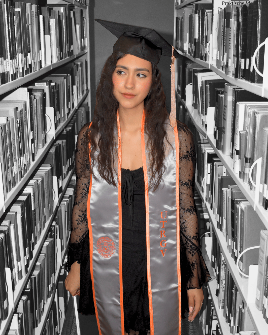 Young woman in graduation cap and gown with honor cords standing in a library between bookshelves, looking to the side.