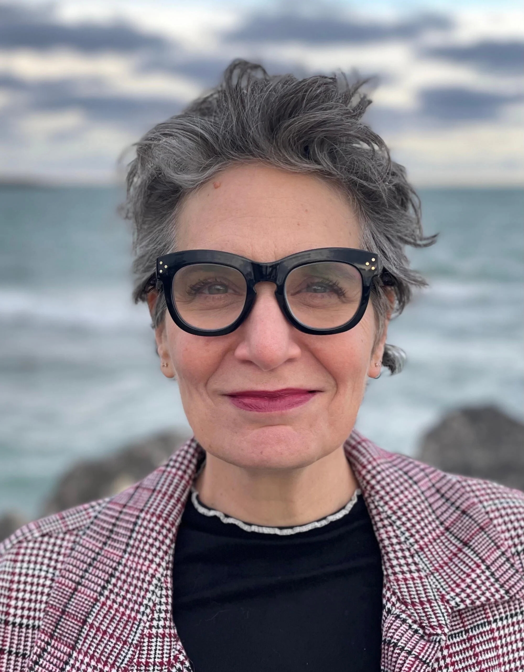 Close-up of a smiling woman with short gray hair, wearing large black glasses, a black shirt with white trim around the neckline, and a red and white checkered jacket, standing outdoors at the beach with rocks and the ocean in the background.