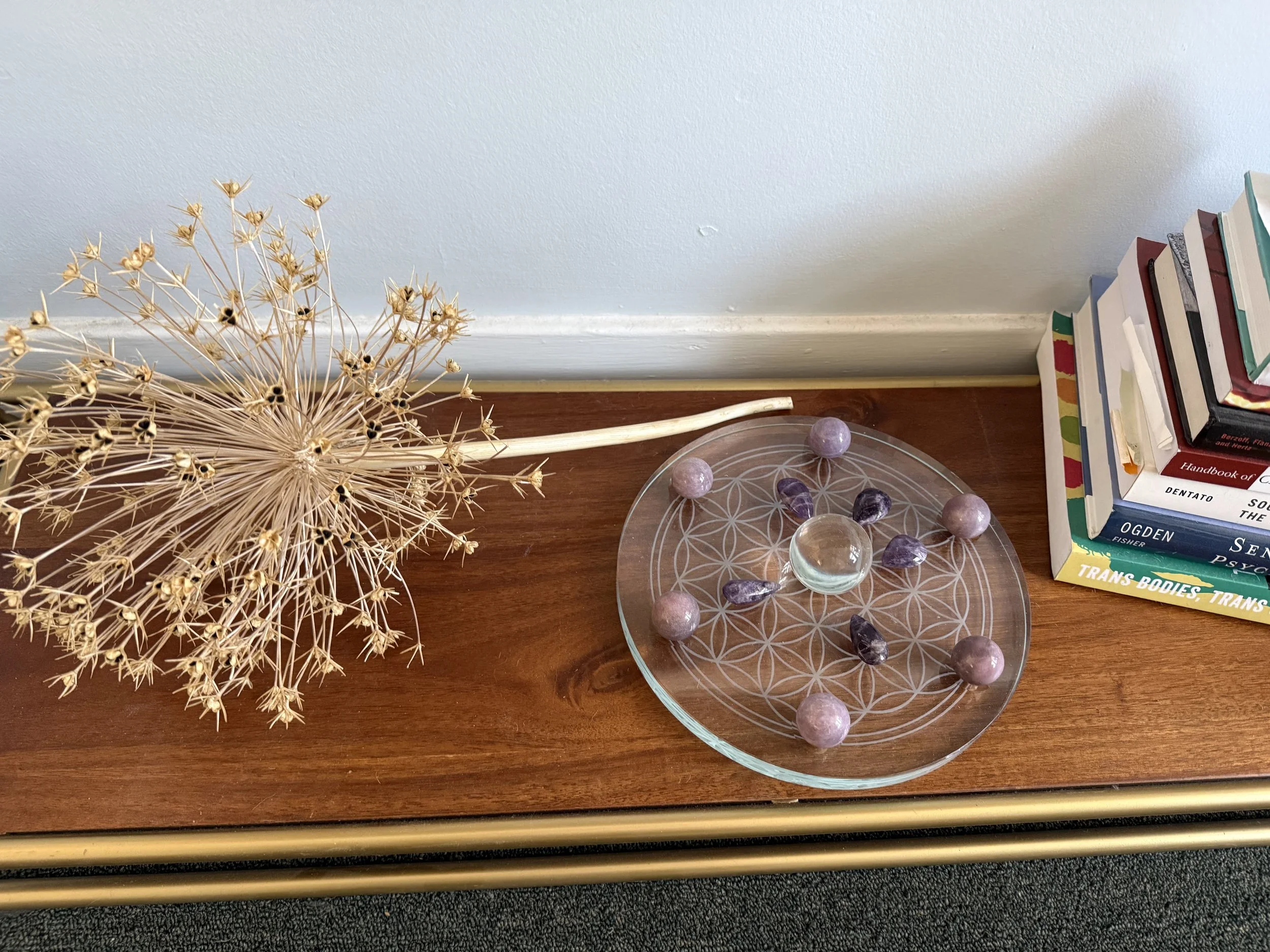 Decorative dried flower arrangement, a glass plate with amethyst stones, and a stack of books on a wooden surface against a pale wall.