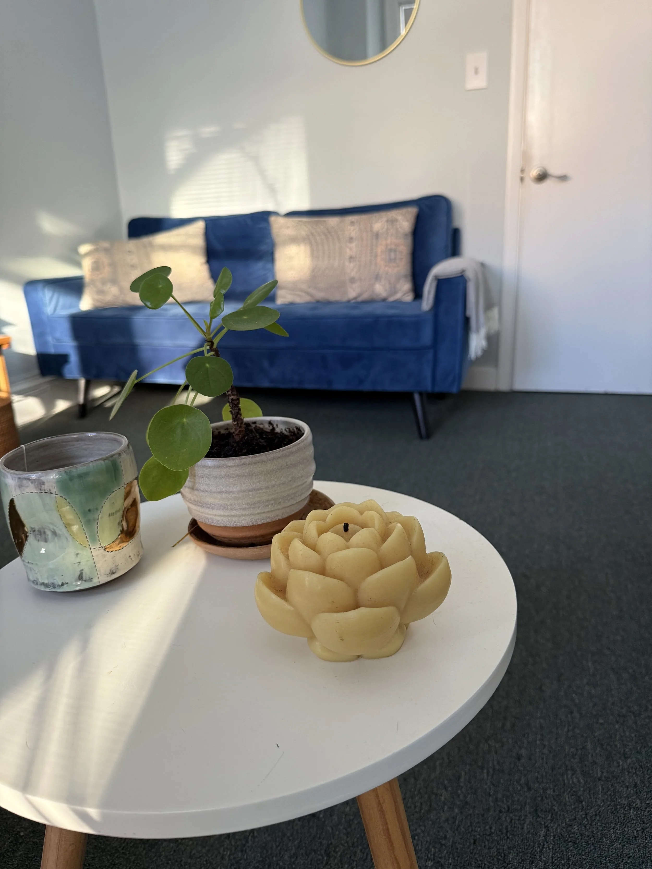 A white circular table with a green potted plant, a decorative candle, and a flower-shaped candle. In the background, there is a blue sofa with pillows, a wall with a round mirror, a door, and a beige towel hanging on the arm of the sofa.