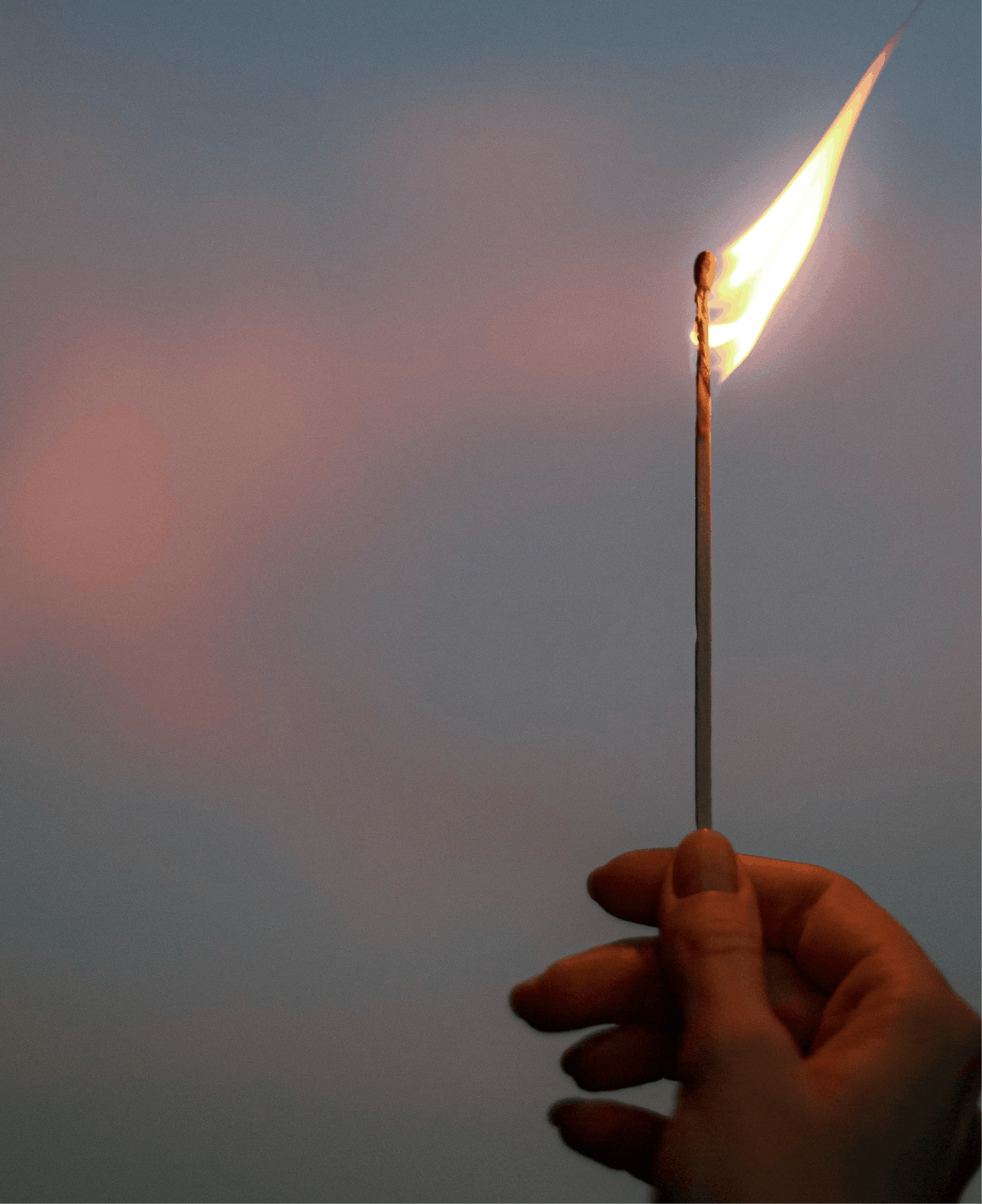 A person holding a lit matchstick that has a small flame at the tip, set against a plain sky background.