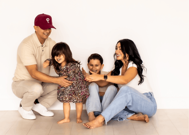 A family of four, including a man, woman, young girl, and young boy, playing and laughing together against a plain white wall. The man is wearing a cap and beige outfit, and the woman and children are casually dressed.